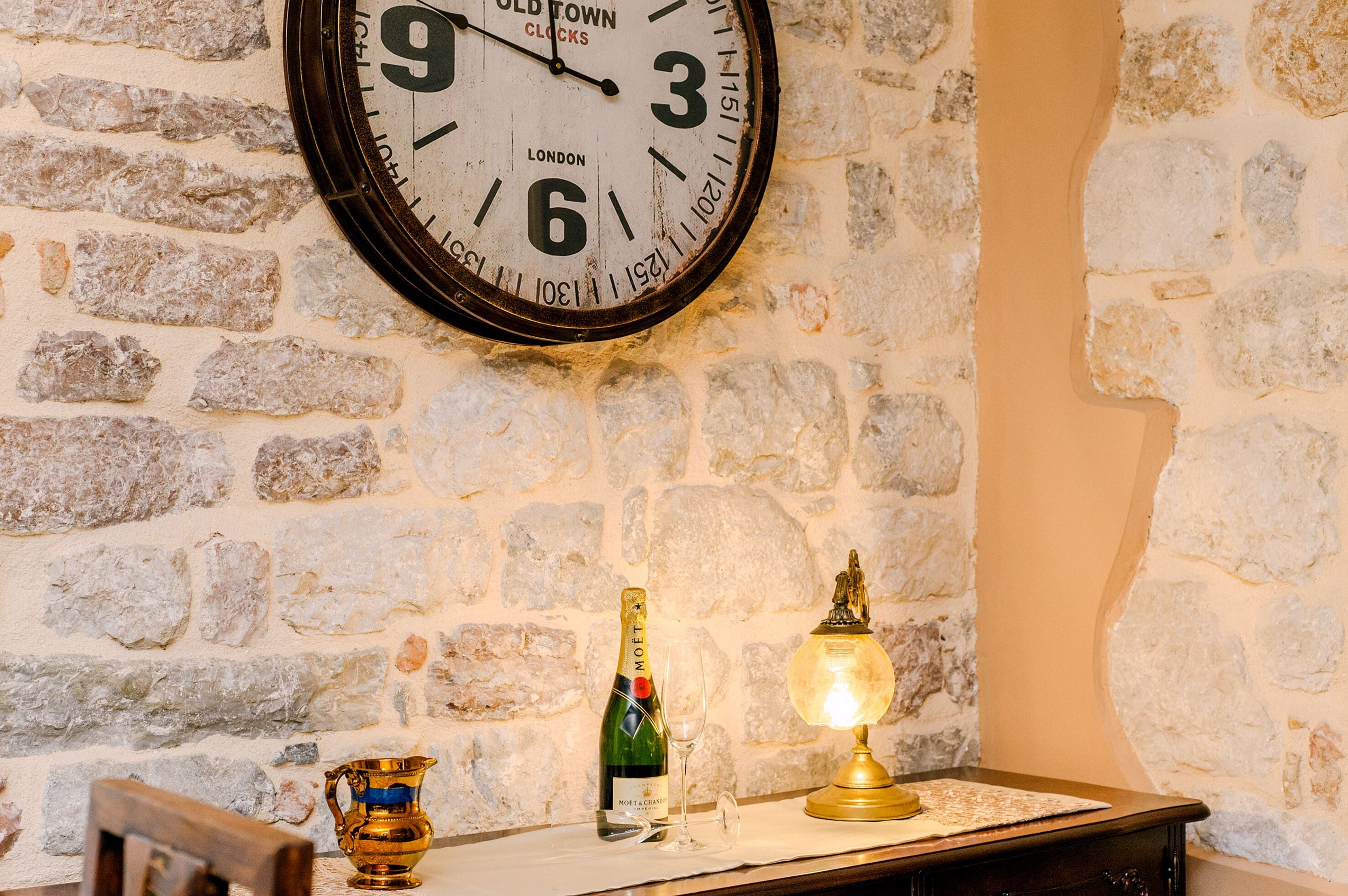 A wall clock, a bottle of champagne, a glass, and a lamp on a table against a stone wall.