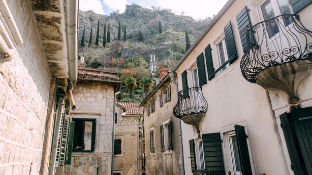 A narrow street with buildings featuring stone walls and green shutters, leading up to a hill with trees and a castle-like structure. - D & Sons Apartments (Kotor)