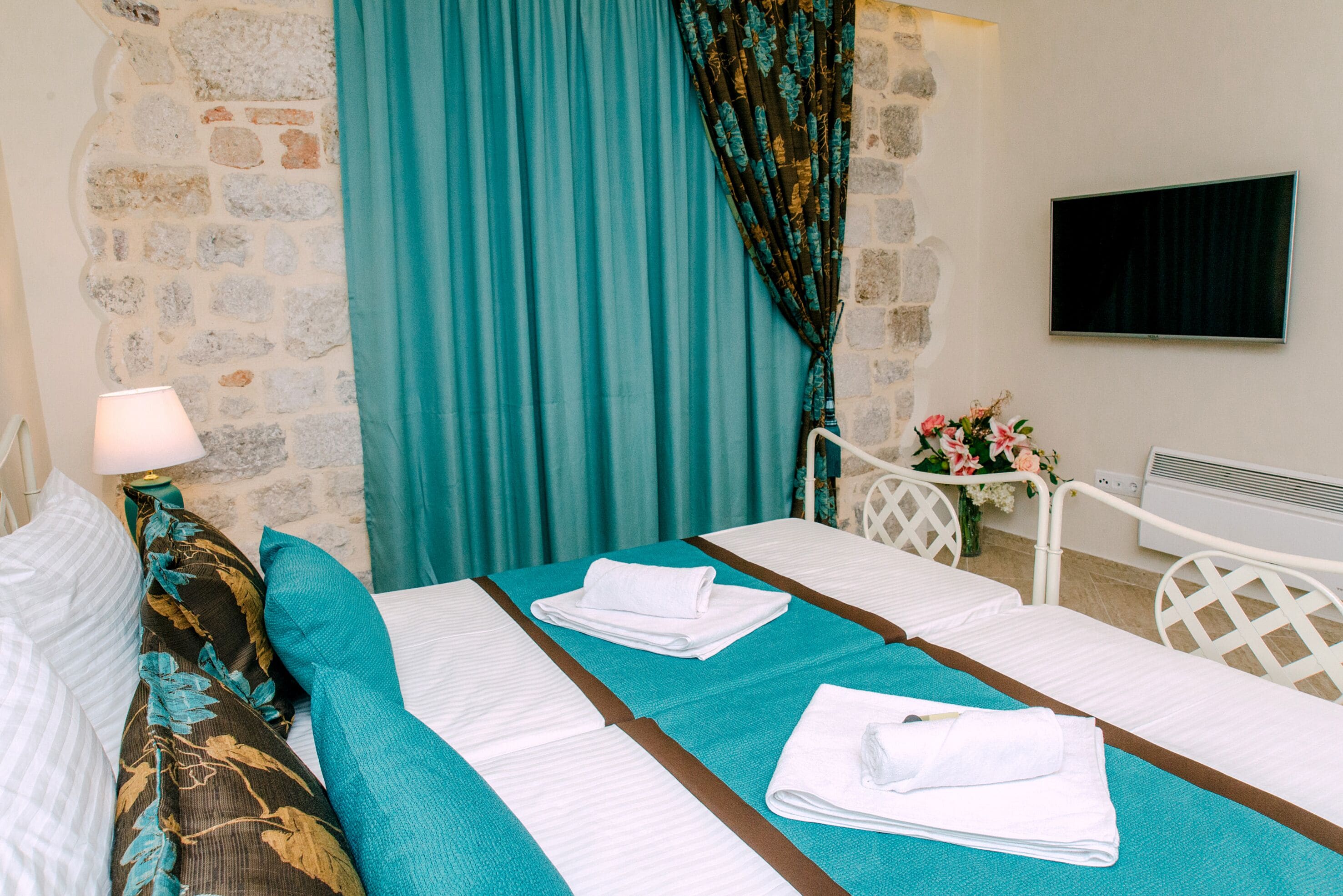 A hotel room with two beds, teal bedding, a stone wall, a TV, and a flower vase.