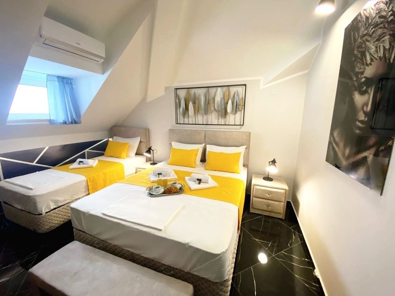 A hotel room with a large bed, two smaller beds, a desk, and a chair. There is a window with curtains, a painting on the wall, and an air conditioning unit.