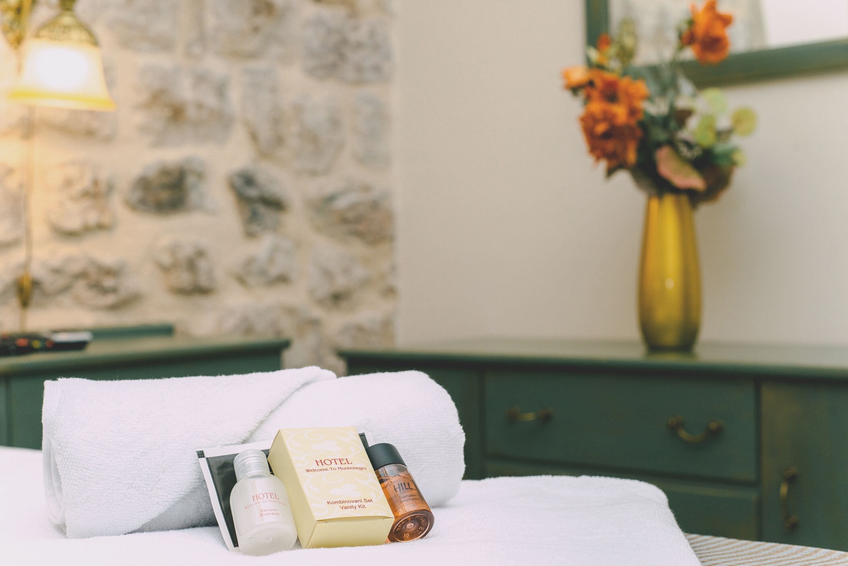 Een net opgemaakt bed met een witte handdoek, fles hotelshampoo en een welkomstpakket op een nachtkastje.