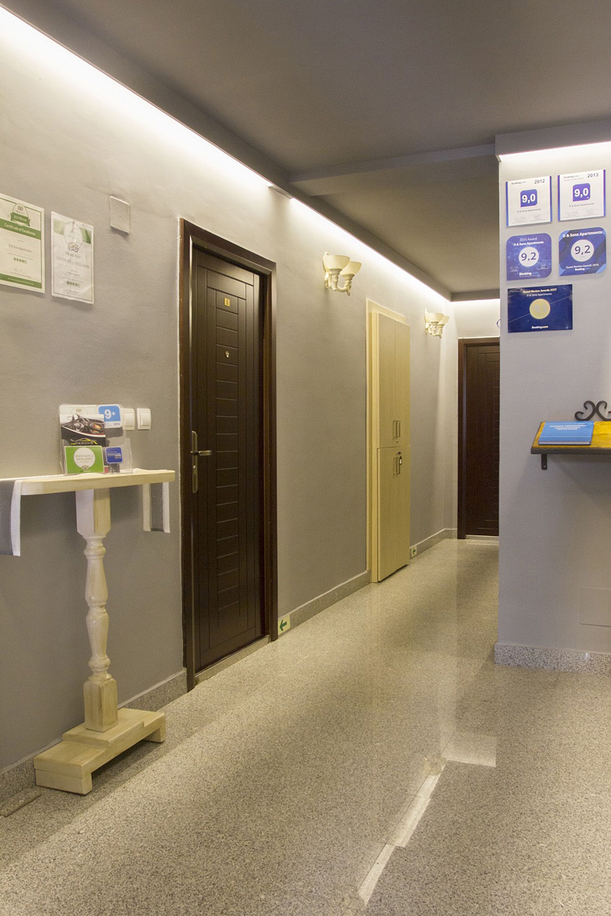Un pasillo de hotel con una puerta de madera, un pedestal blanco y un estante de pared con certificados.