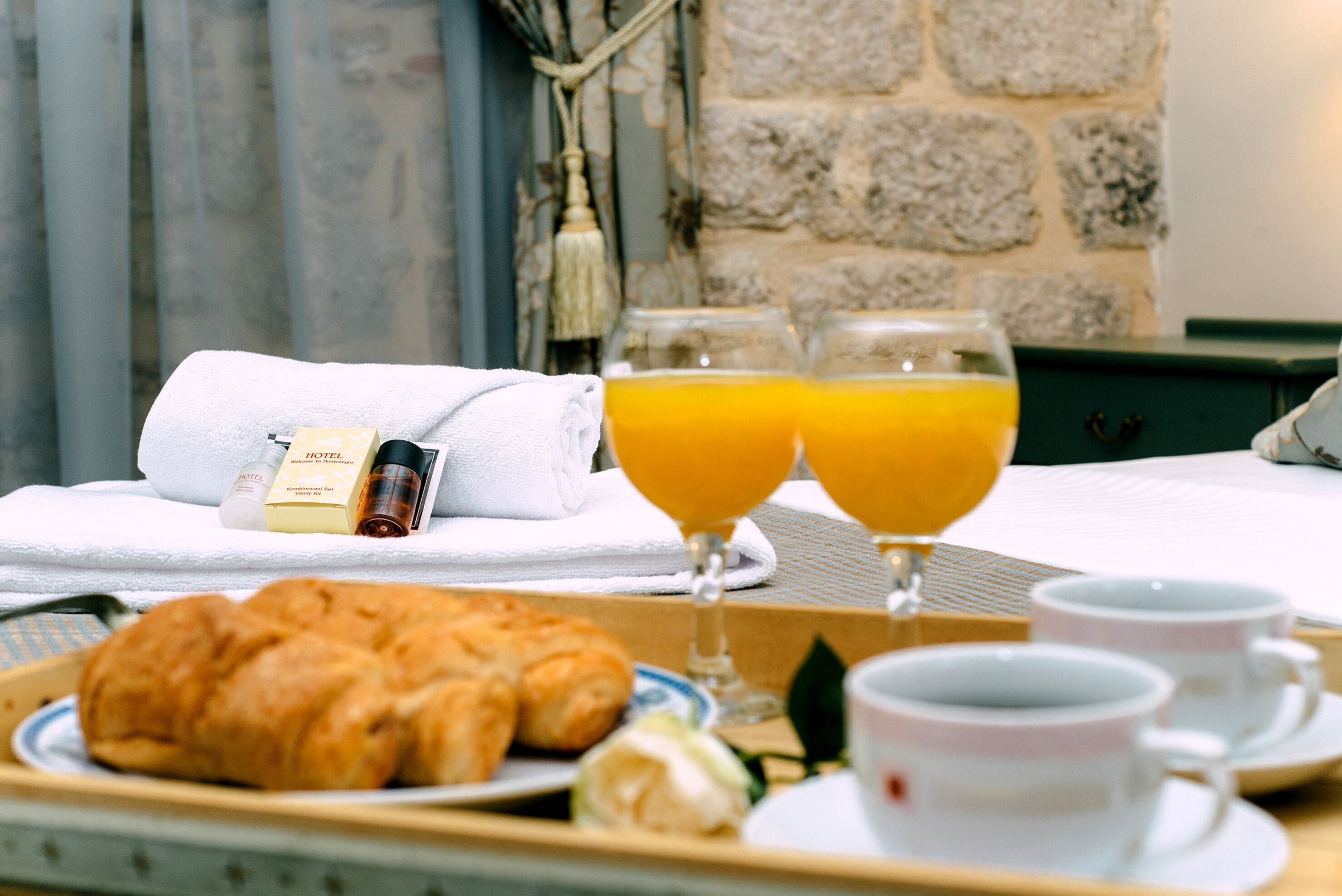 A breakfast tray with croissants, juice, and coffee cups, a towel, and a hotel amenity bottle on a bed in a room with stone walls and a window.