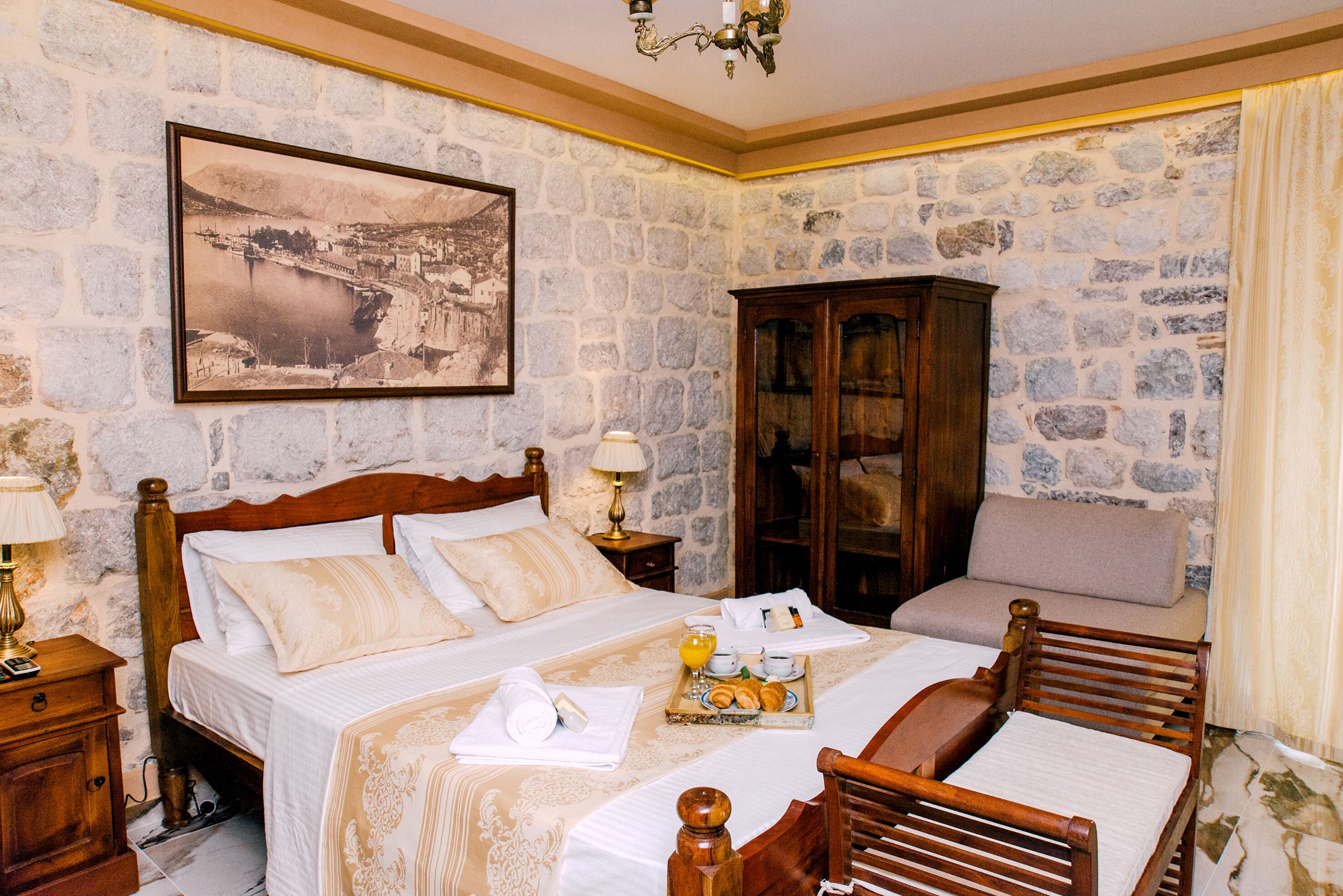 A hotel room with stone walls, a large bed with a wooden headboard, a wooden cabinet, and a framed picture on the wall.