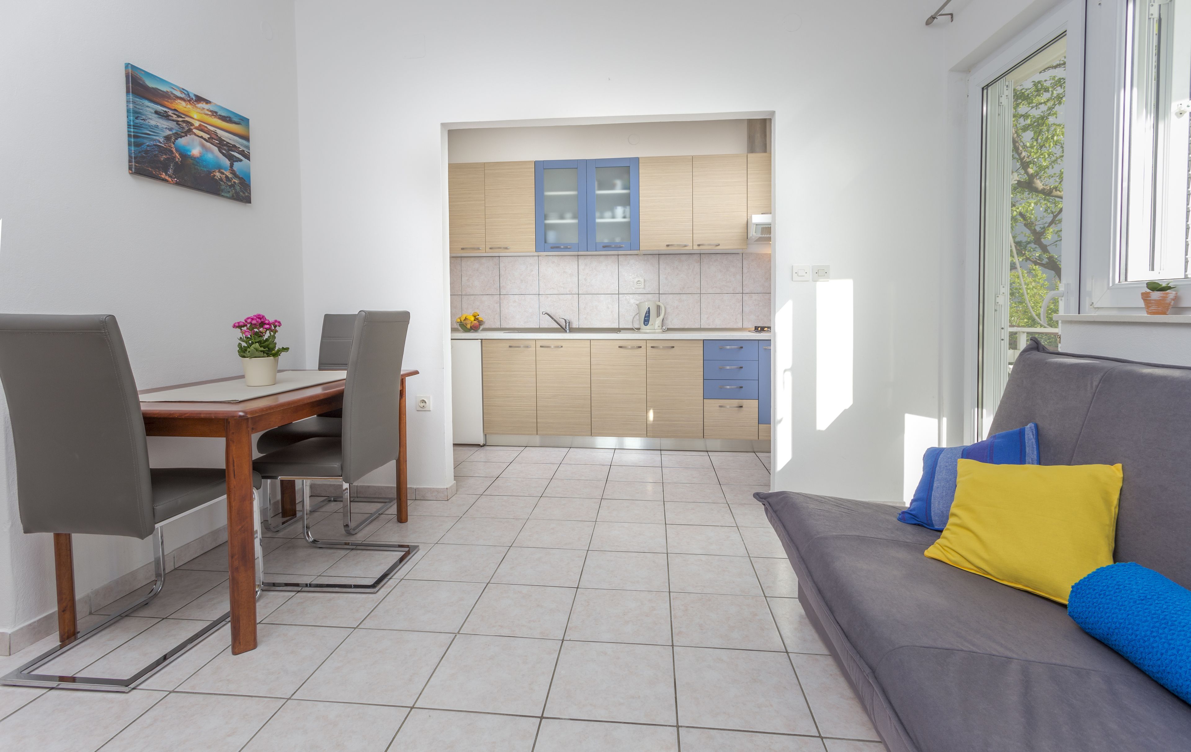 Una cocina moderna con armarios azules, una zona de comedor con una mesa y sillas de madera, y un sofá gris con cojines coloridos.