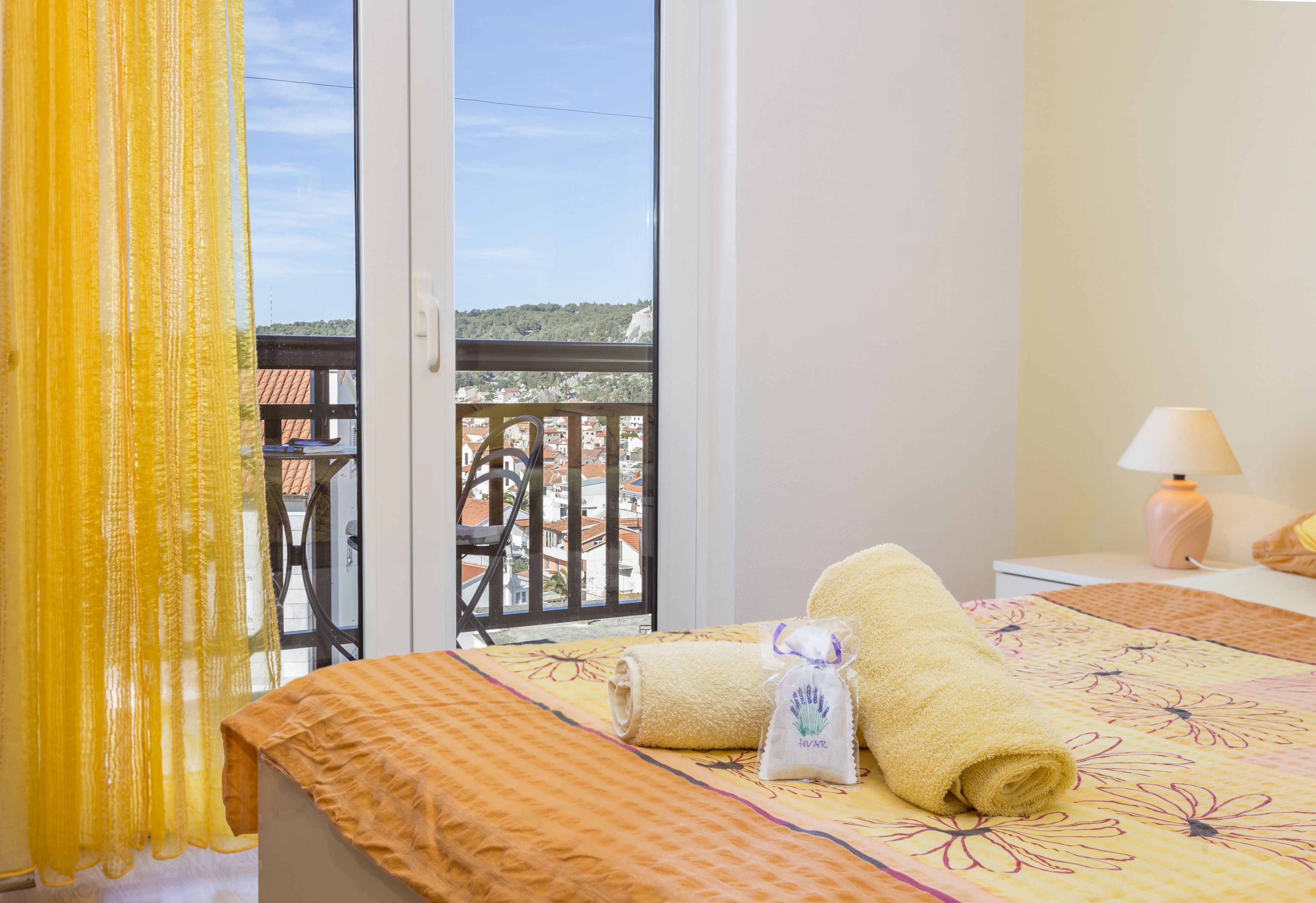 A hotel room with a bed, a window with a view of buildings, a balcony, and a lamp on the nightstand.