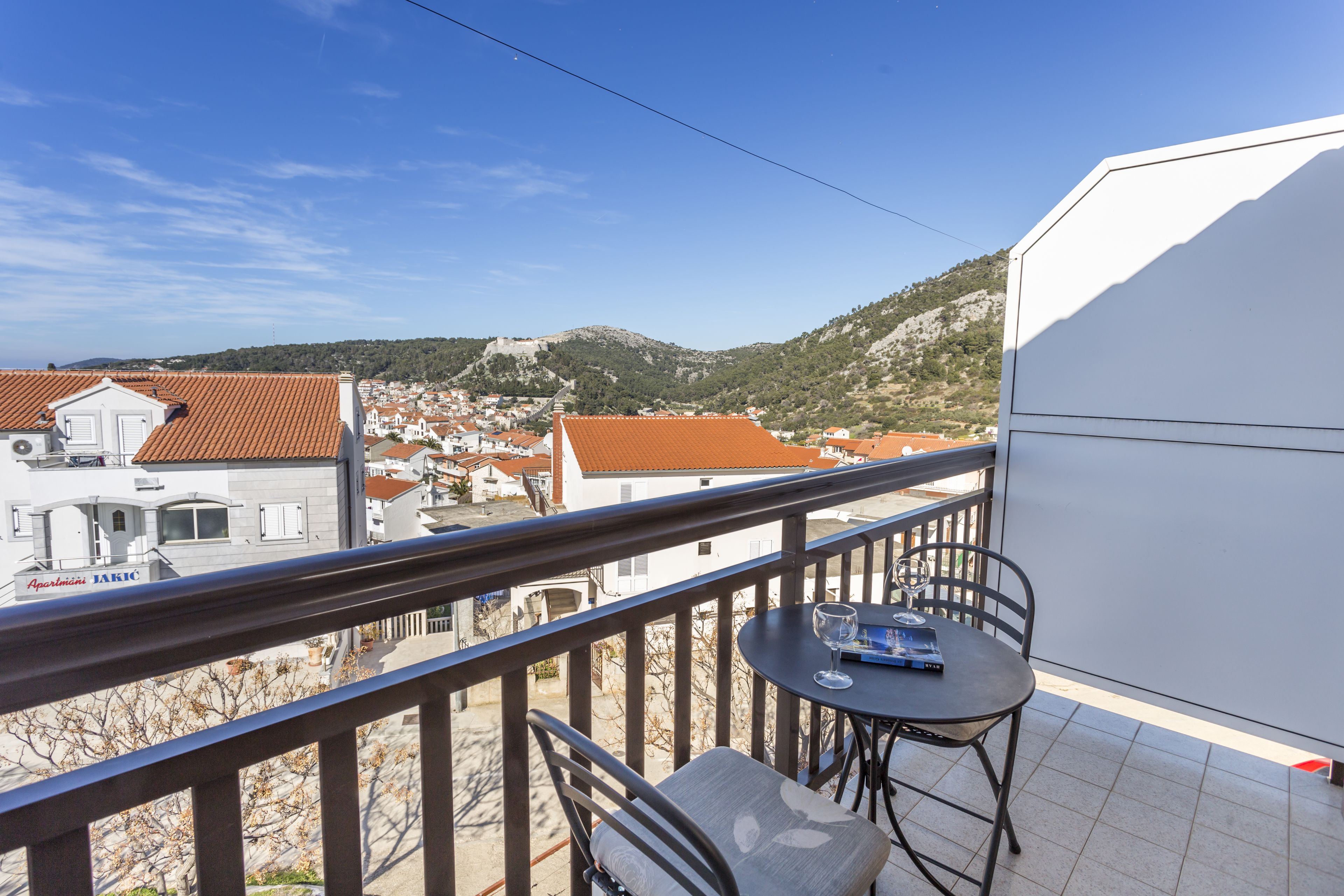 A balcony with a table and chairs, overlooking a town with red-roofed buildings and a hill in the background.