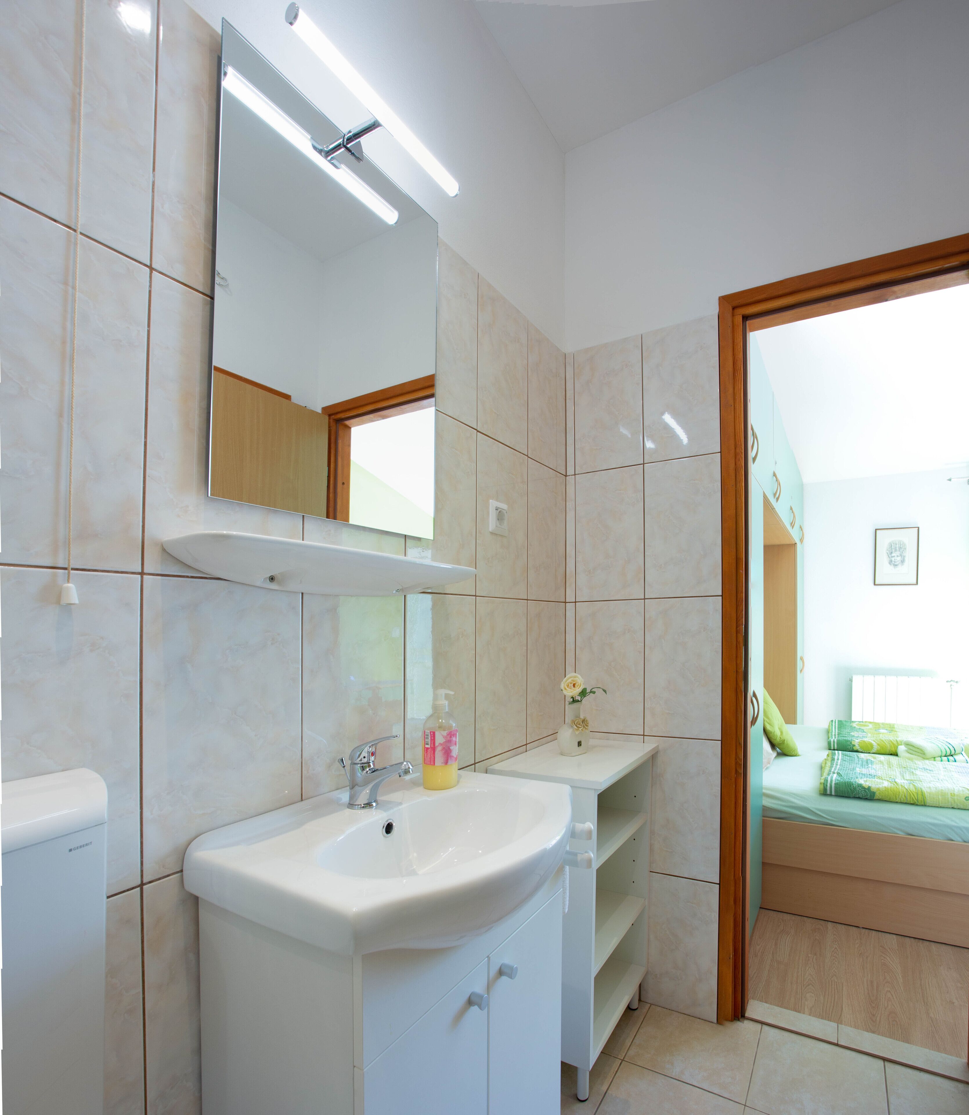 A bathroom with a white sink, mirror, and toilet. There is a small shelf with a bottle and a vase with flowers. The room has beige tiles and a wooden door leading to a bedroom.