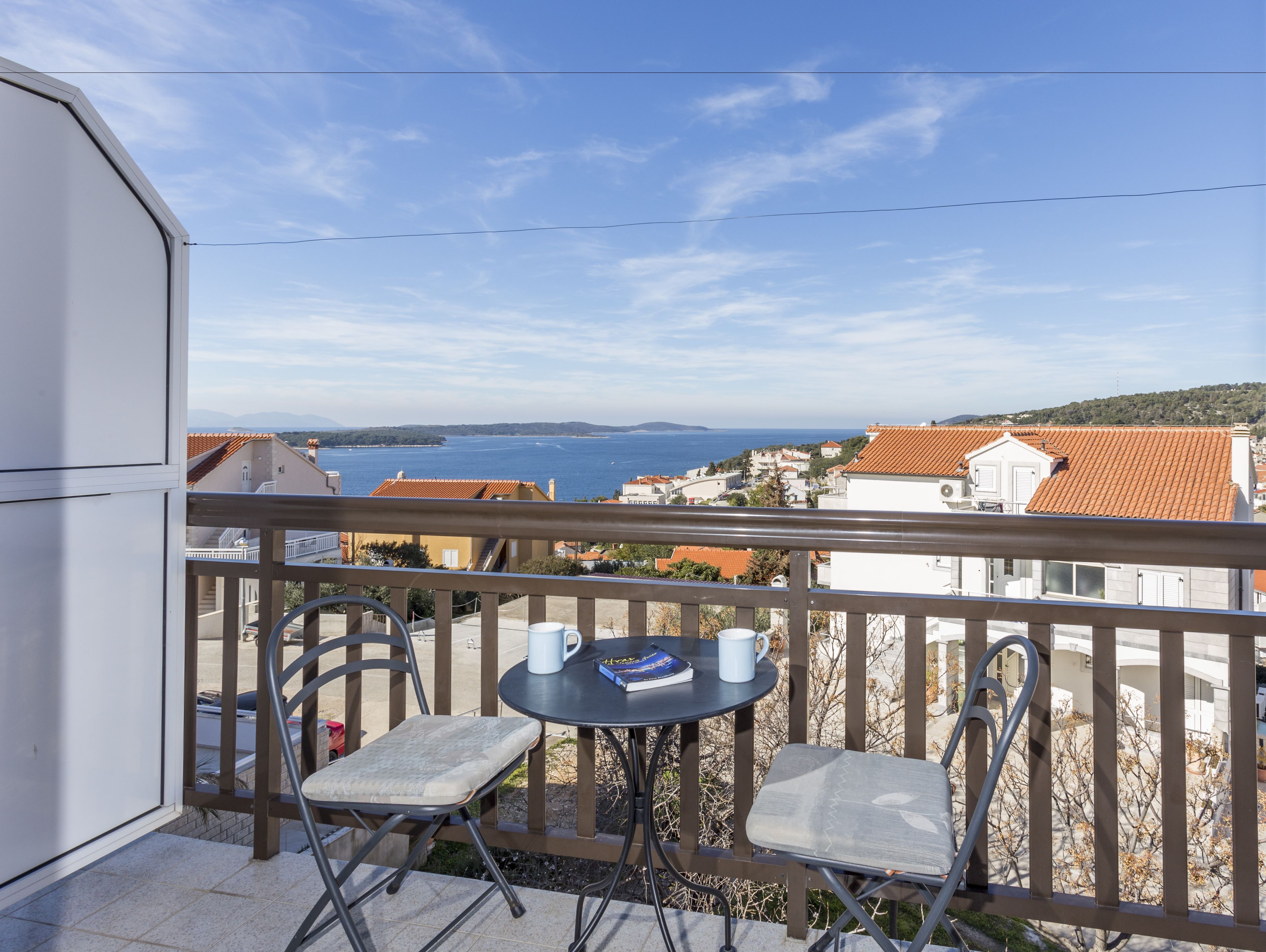 Ein Balkon mit zwei Stühlen und einem kleinen Tisch, mit Blick auf eine Küstenstadt mit terrakottafarbenen Dächern und freiem Blick auf das Meer.