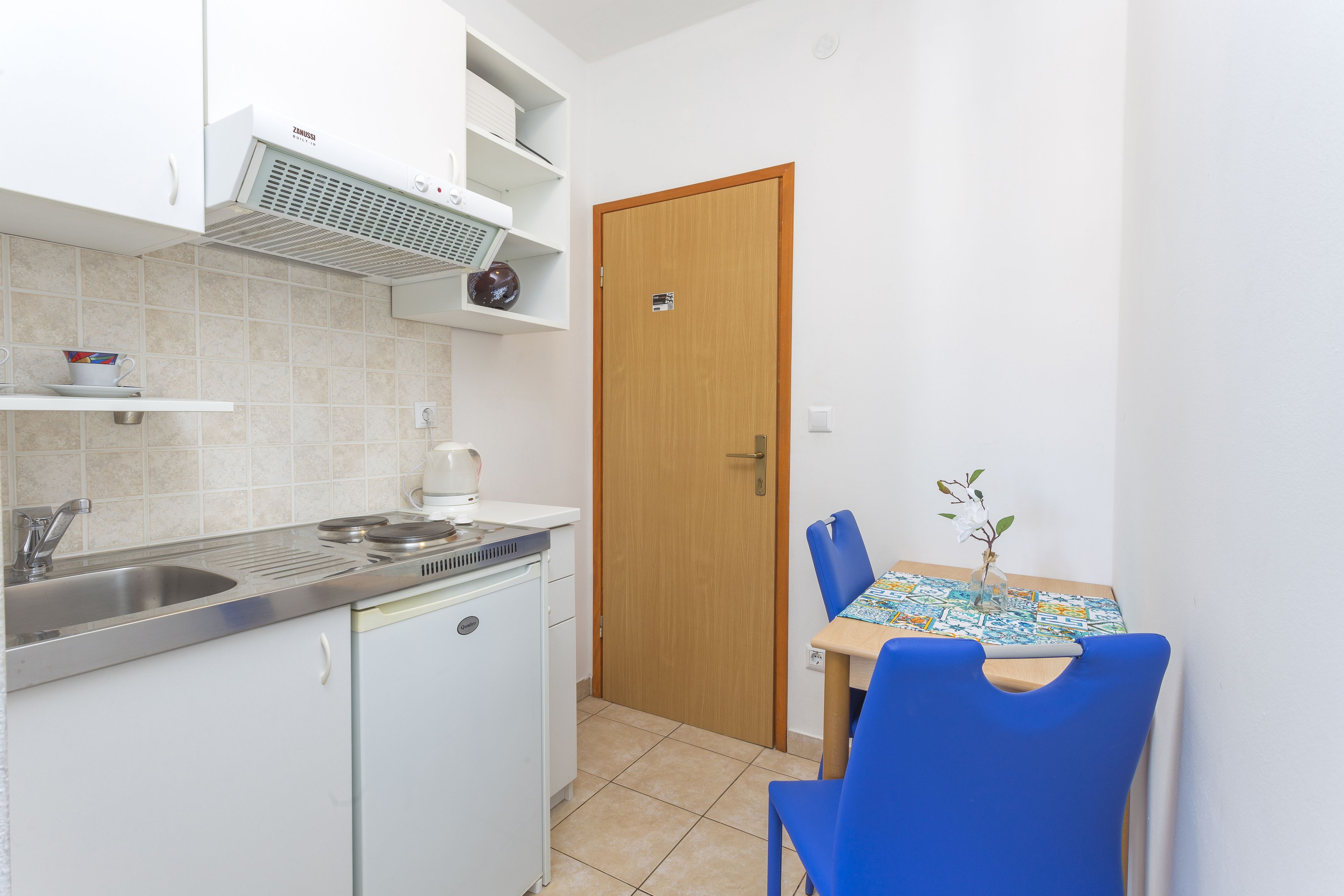 A compact kitchen with a sink, stove, and white cabinets. A wooden door and a small dining table with blue chairs are visible.