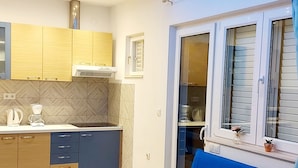 A modern living room with a blue sofa and yellow pillows, a kitchen with wooden cabinets and a built-in stove, and a window with blinds. - Apartmani Ivanovic (Hvar)