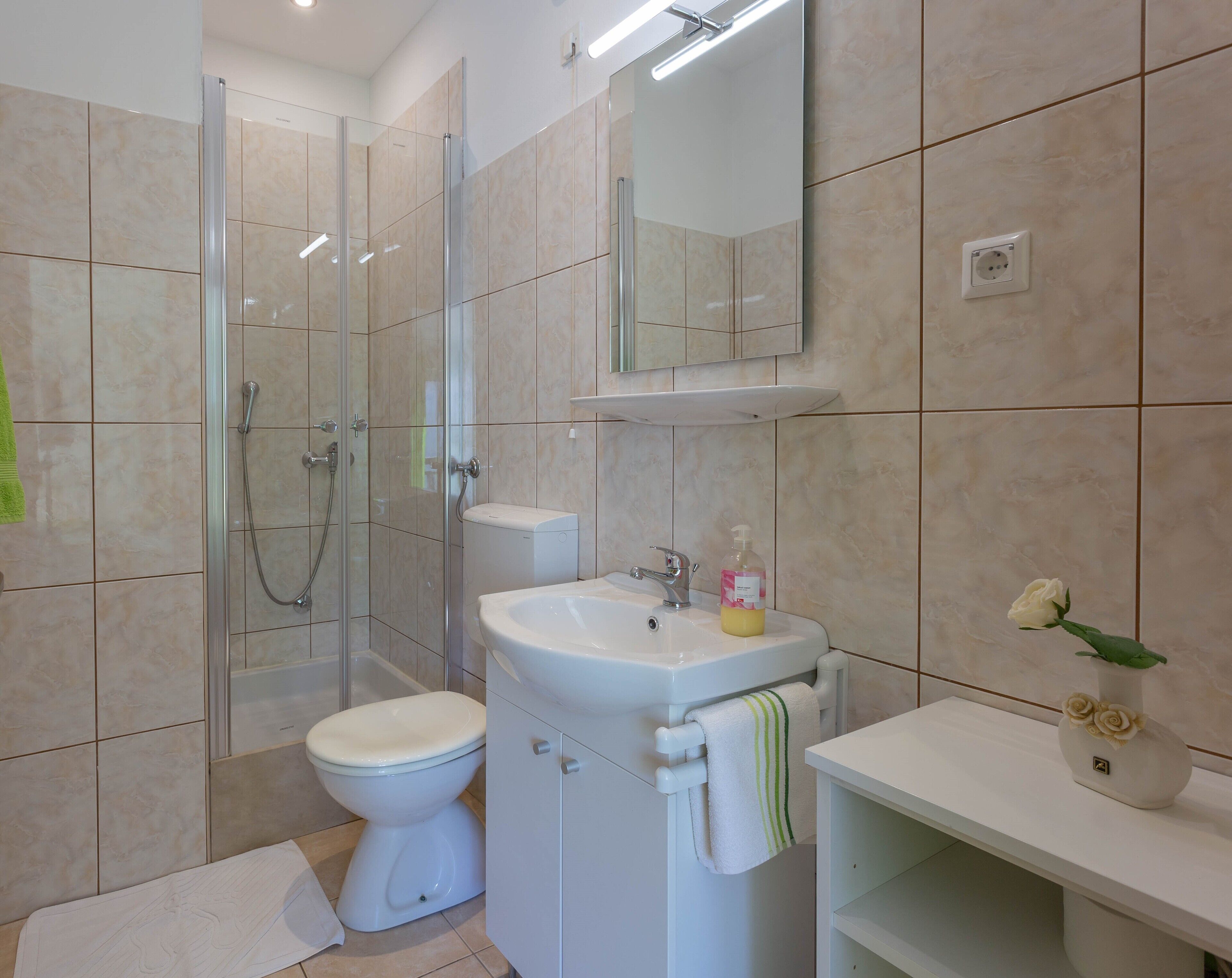 Un baño moderno con ducha de vidrio, inodoro, lavamanos con espejo y un florero con flores sobre la encimera.