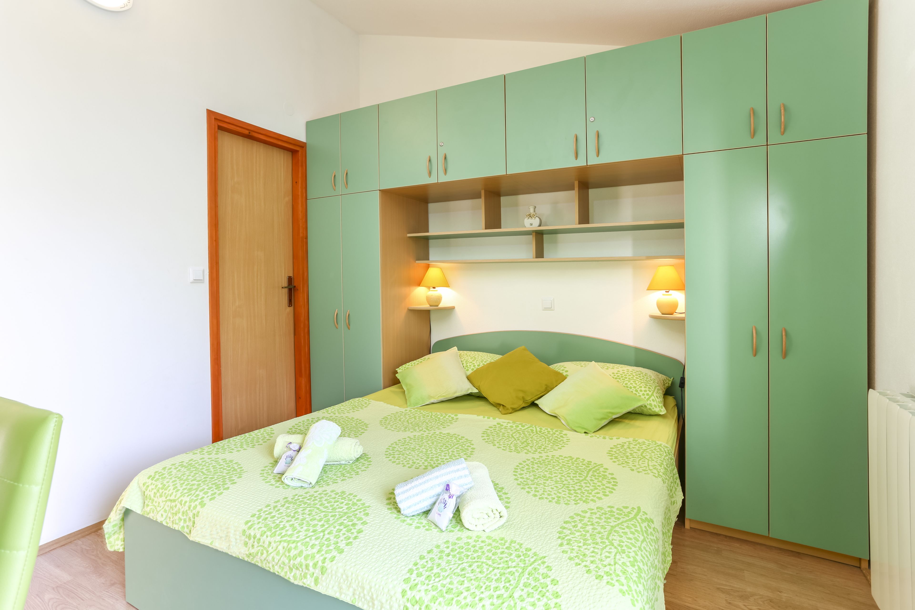 A neatly made bed with a green patterned bedspread, a wooden door, and green cabinets with shelves.
