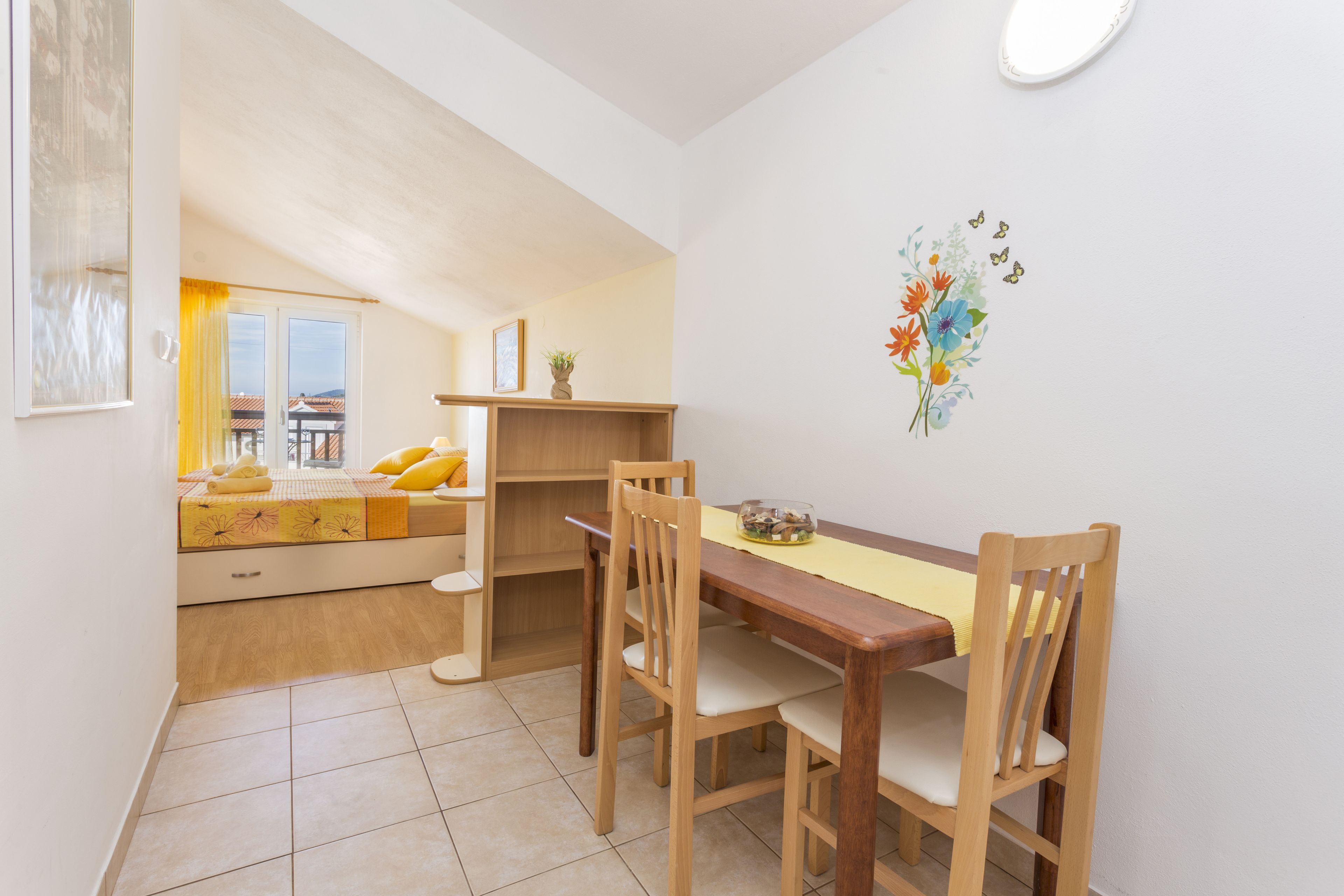 A compact room with a bed, a small table, and a chair. There is a window with curtains, a wall-mounted shelf, and a wall decoration.
