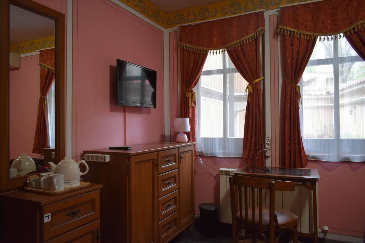 Une pièce aux murs roses, avec une commode en bois, un téléviseur, un bureau avec une chaise et une fenêtre avec des rideaux.