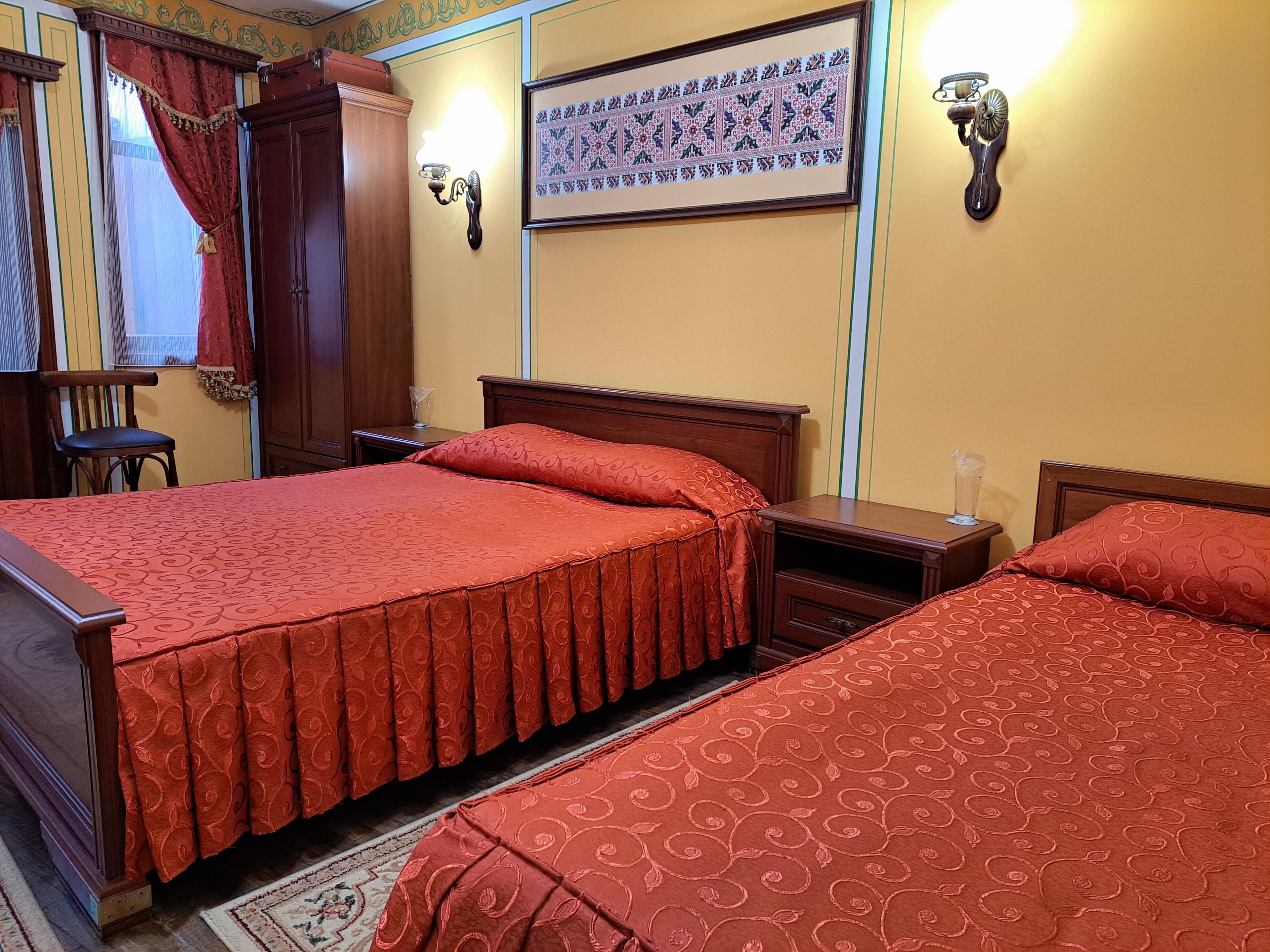 A hotel room with two beds, a wooden wardrobe, a nightstand, and a wall-mounted lamp.