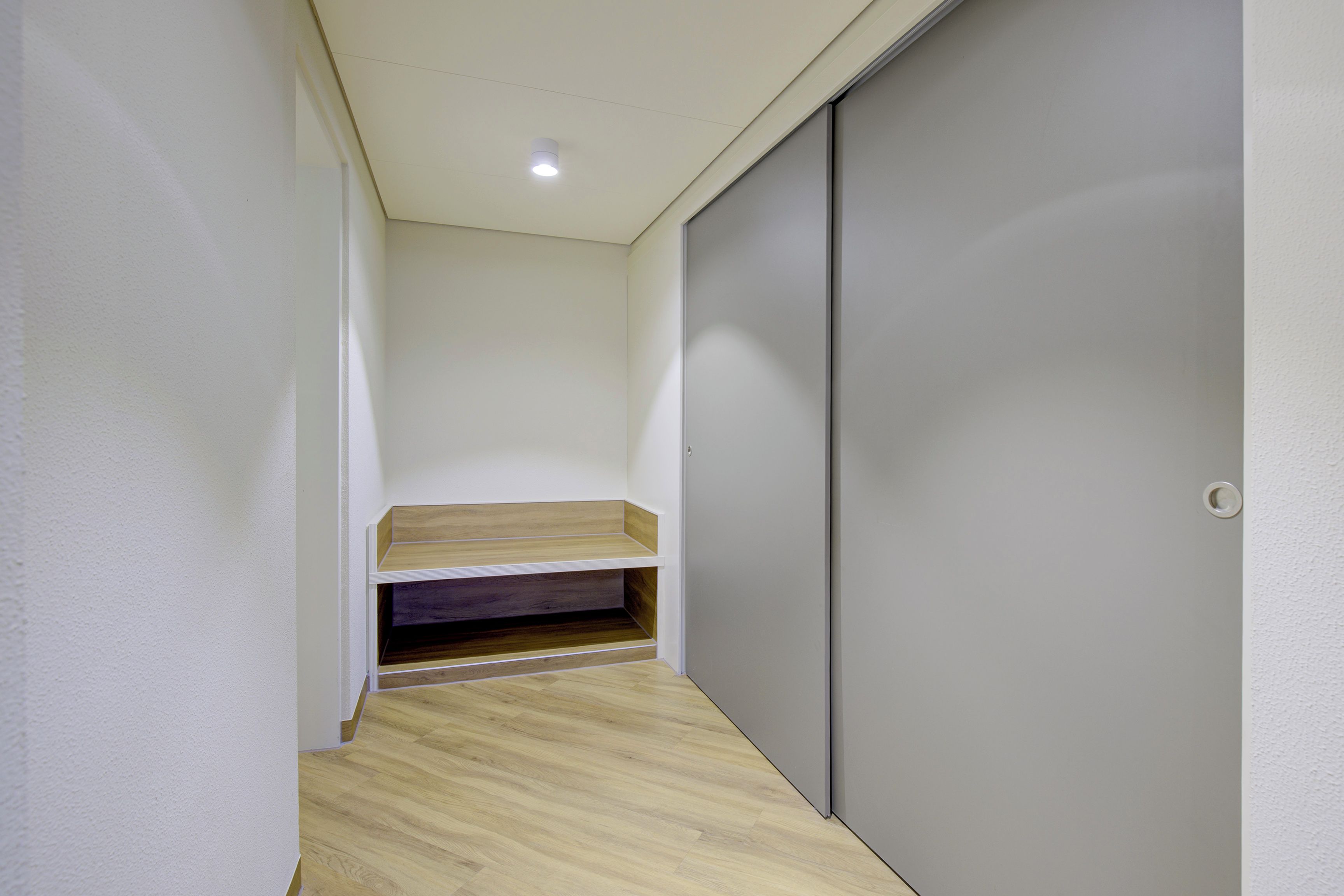 A narrow hallway with a wooden bench, a grey sliding door, and a recessed ceiling light.