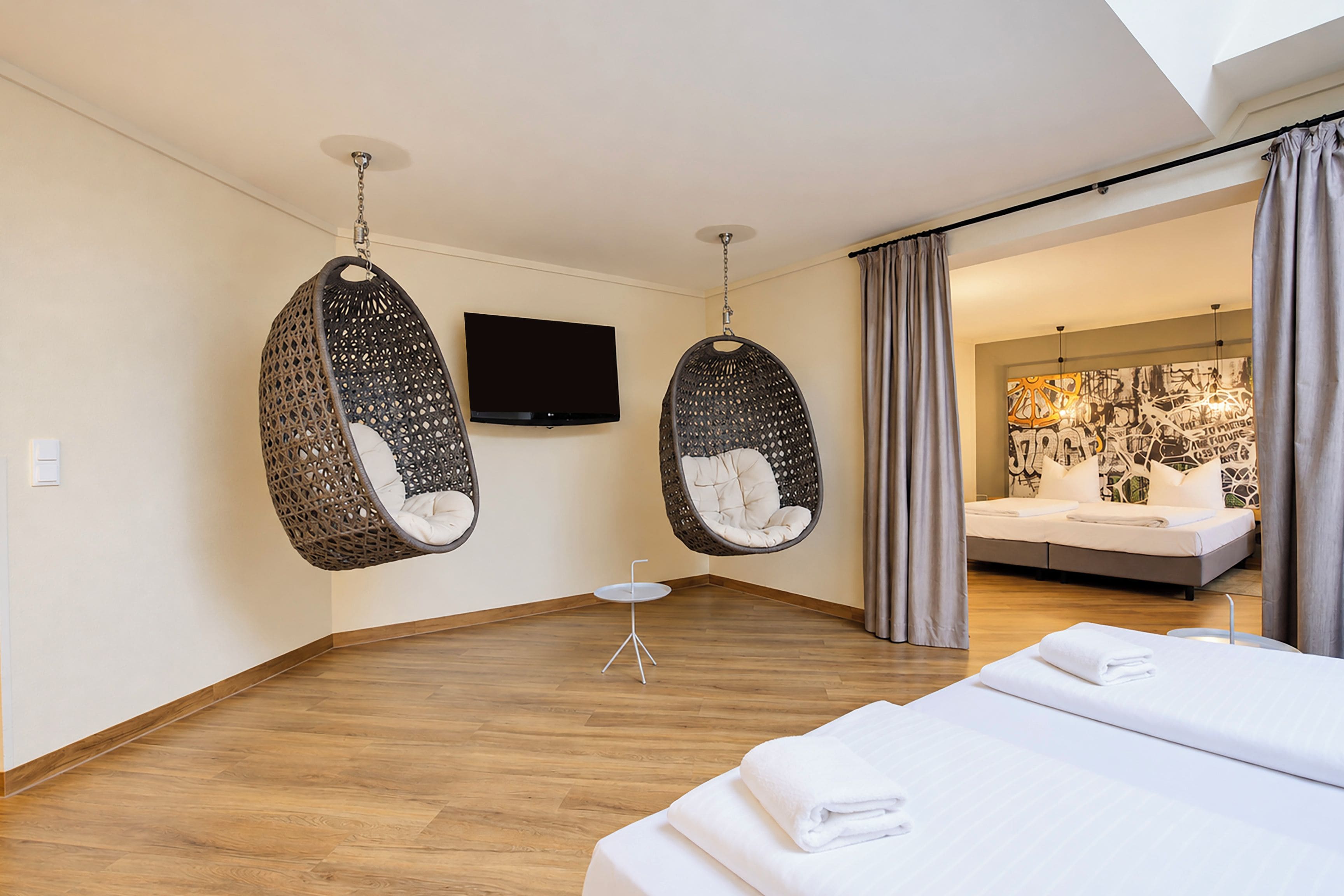 Una habitación de hotel moderna con suelo de madera, dos sillas colgantes, un televisor de pantalla plana, una mesita redonda y una cama con toallas ordenadas.