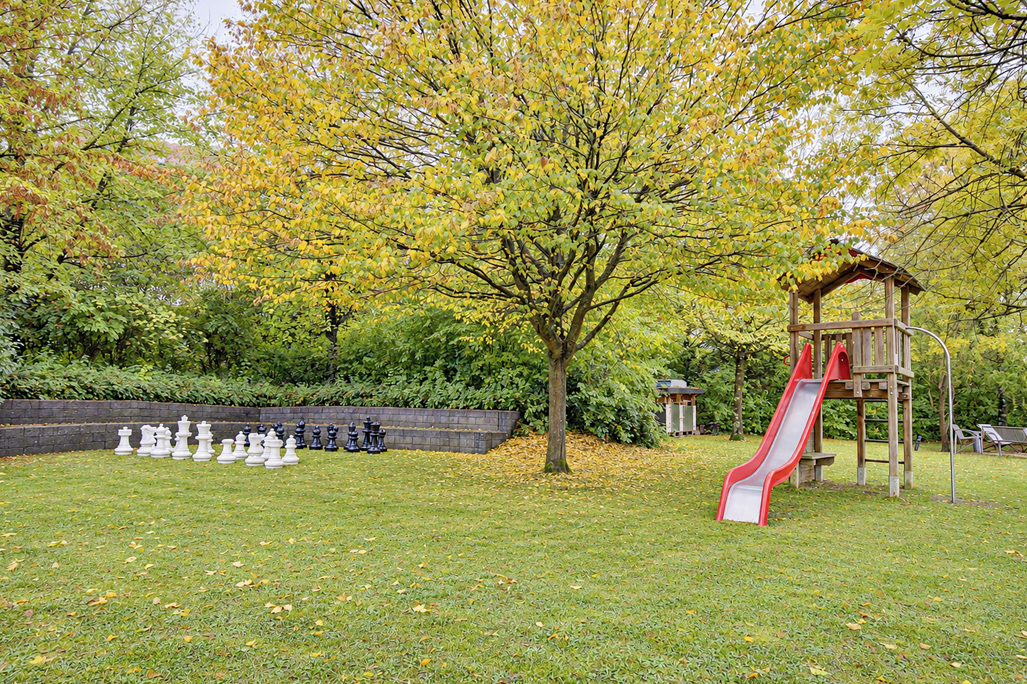 Ein Spielplatz mit Rutsche und Klettergerüst, umgeben von Bäumen und einem Schachbrett auf der Wiese.