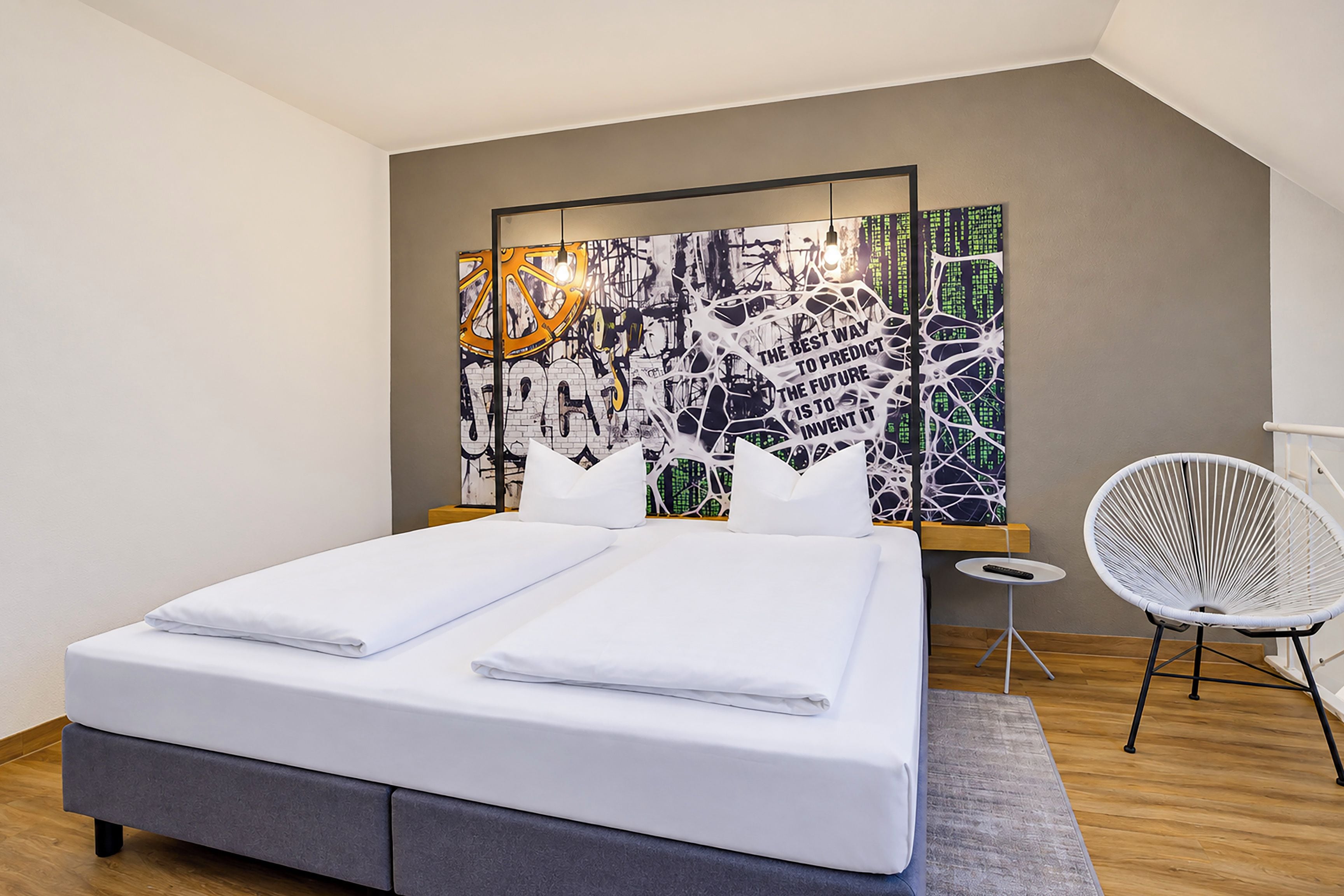 Ein modernes Schlafzimmer mit einem ordentlich bezogenen Bett, einem Wandbild, einem kleinen Nachttisch und einem weißen Stuhl.