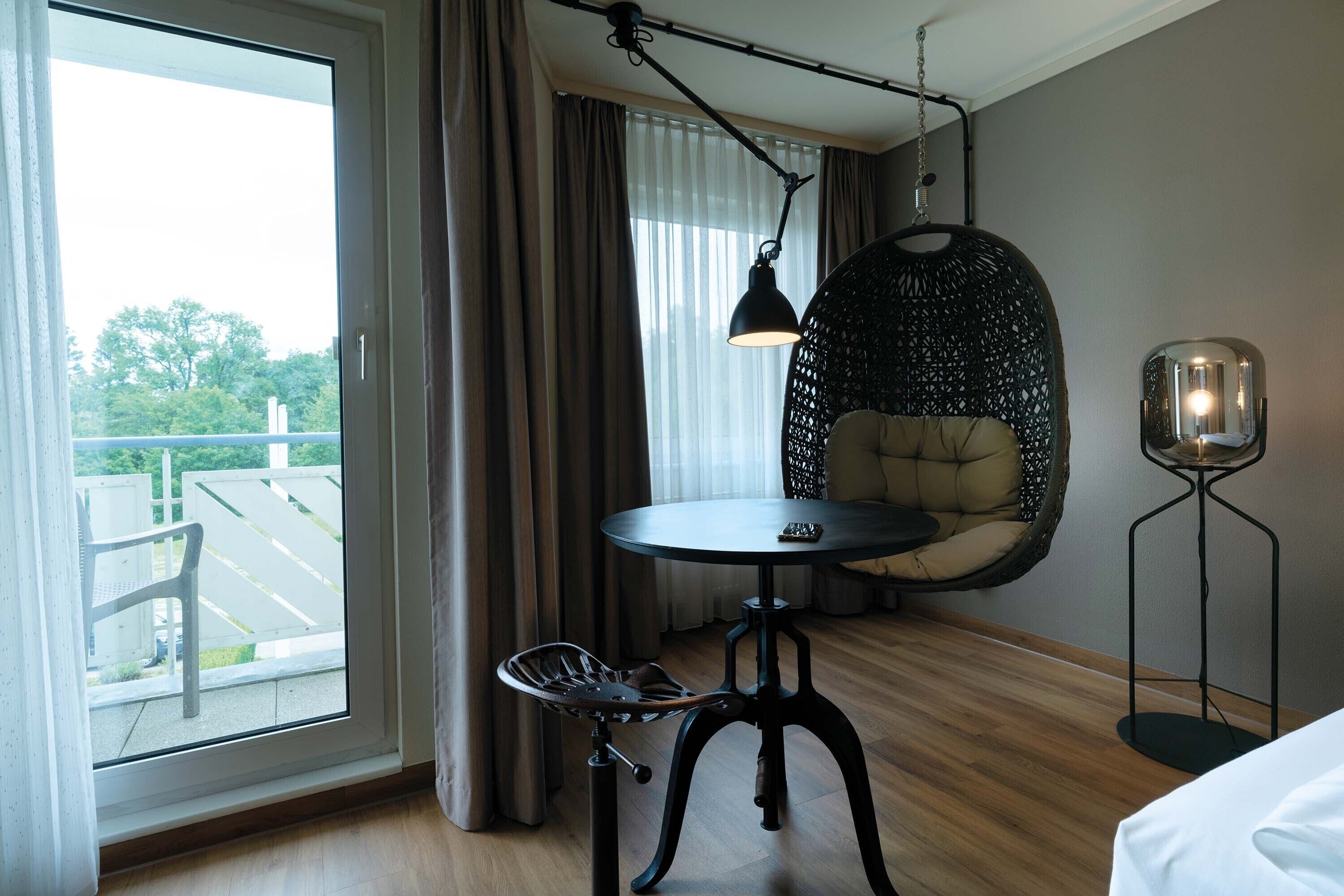 Moderni hotellihuone, jossa on suuri ikkuna, pyöreä pöytä, riipputuoli ja lattiavalaisin.