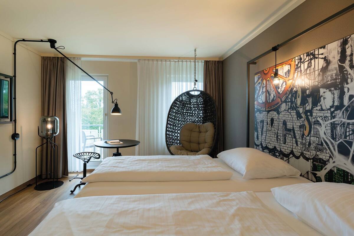 A hotel room with two beds, a hanging chair, a floor lamp, and a large abstract painting on the wall.