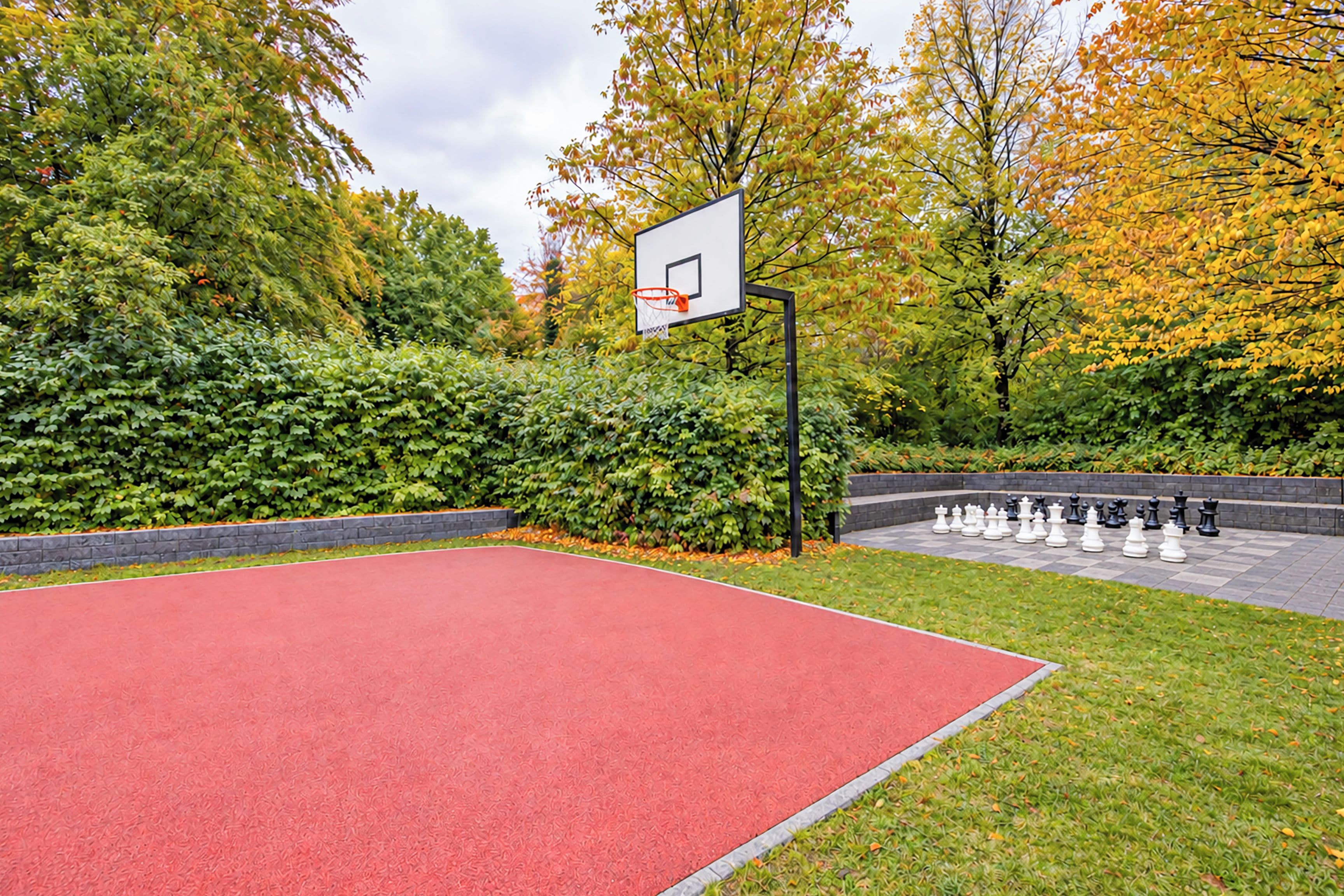 Una cancha de baloncesto al aire libre con superficie roja, rodeada de árboles y un tablero de ajedrez.