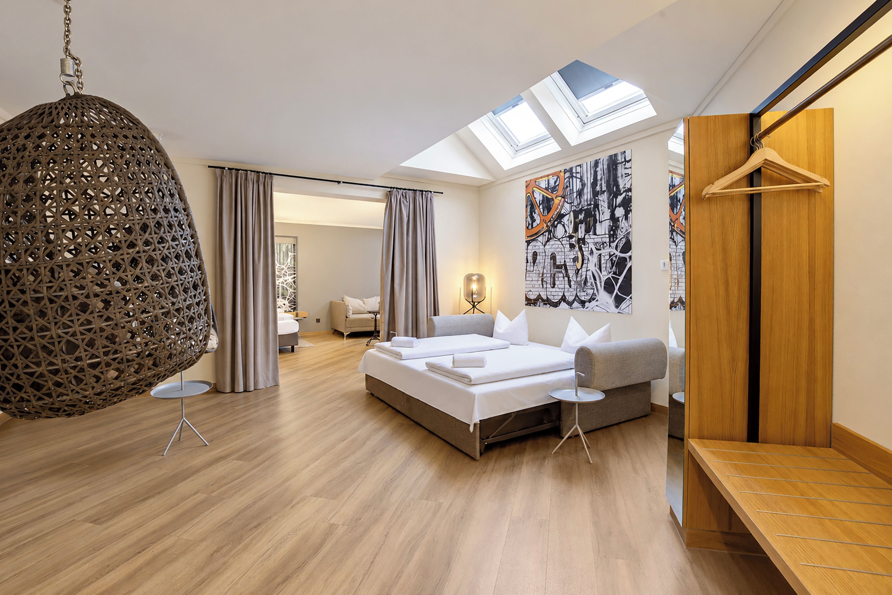 Une chambre d’hôtel moderne dotée d’une lucarne, d’un grand lit, d’un coin salon et d’un mur orné d’œuvres d’art abstraites.