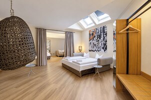 Quarto de hotel moderno com claraboia, uma cama grande, área de estar e parede com arte abstrata.