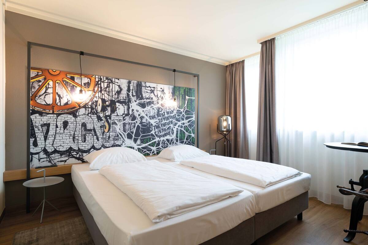 A hotel room with a large bed, a small table, a wall art piece, and a window with curtains.