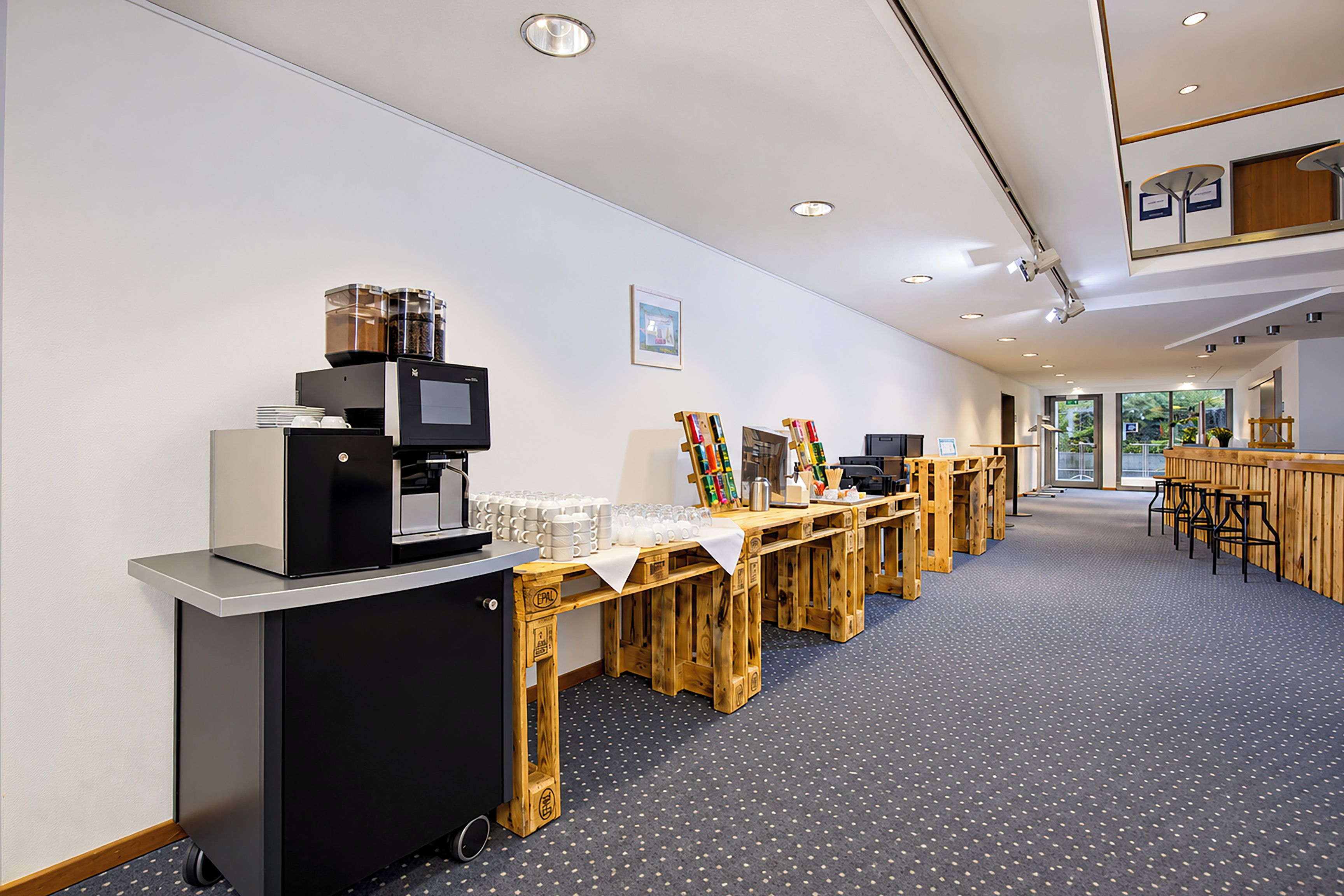 Moderni hotellin aula, jossa on kahvipiste, puupöytiä ja näkymä ulos.