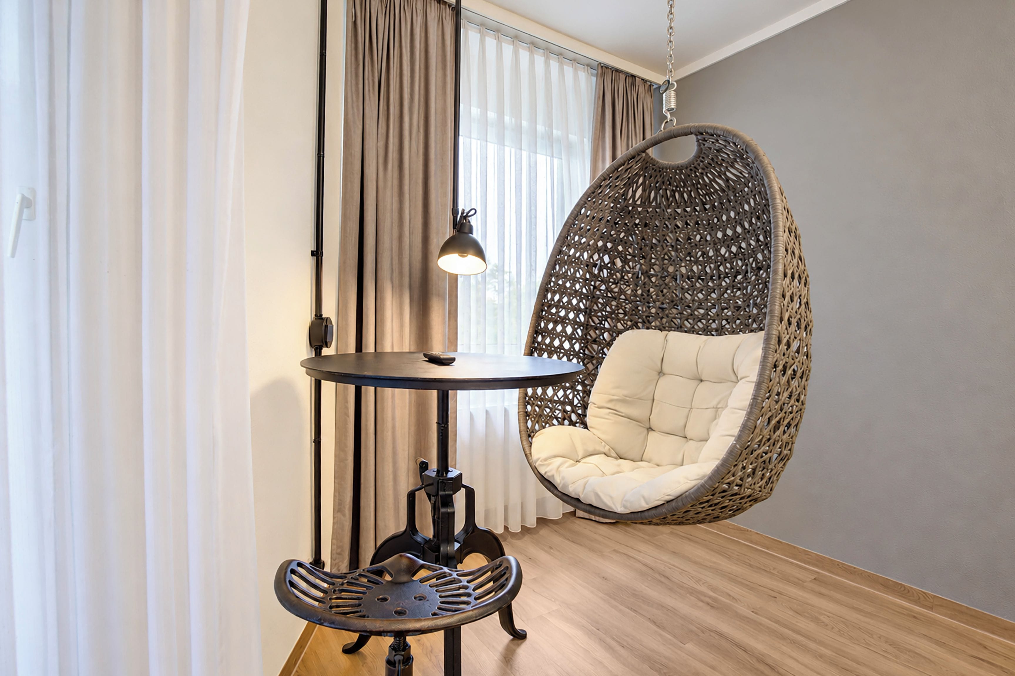 A room with a hanging chair, a small table with a lamp, and a window with curtains.