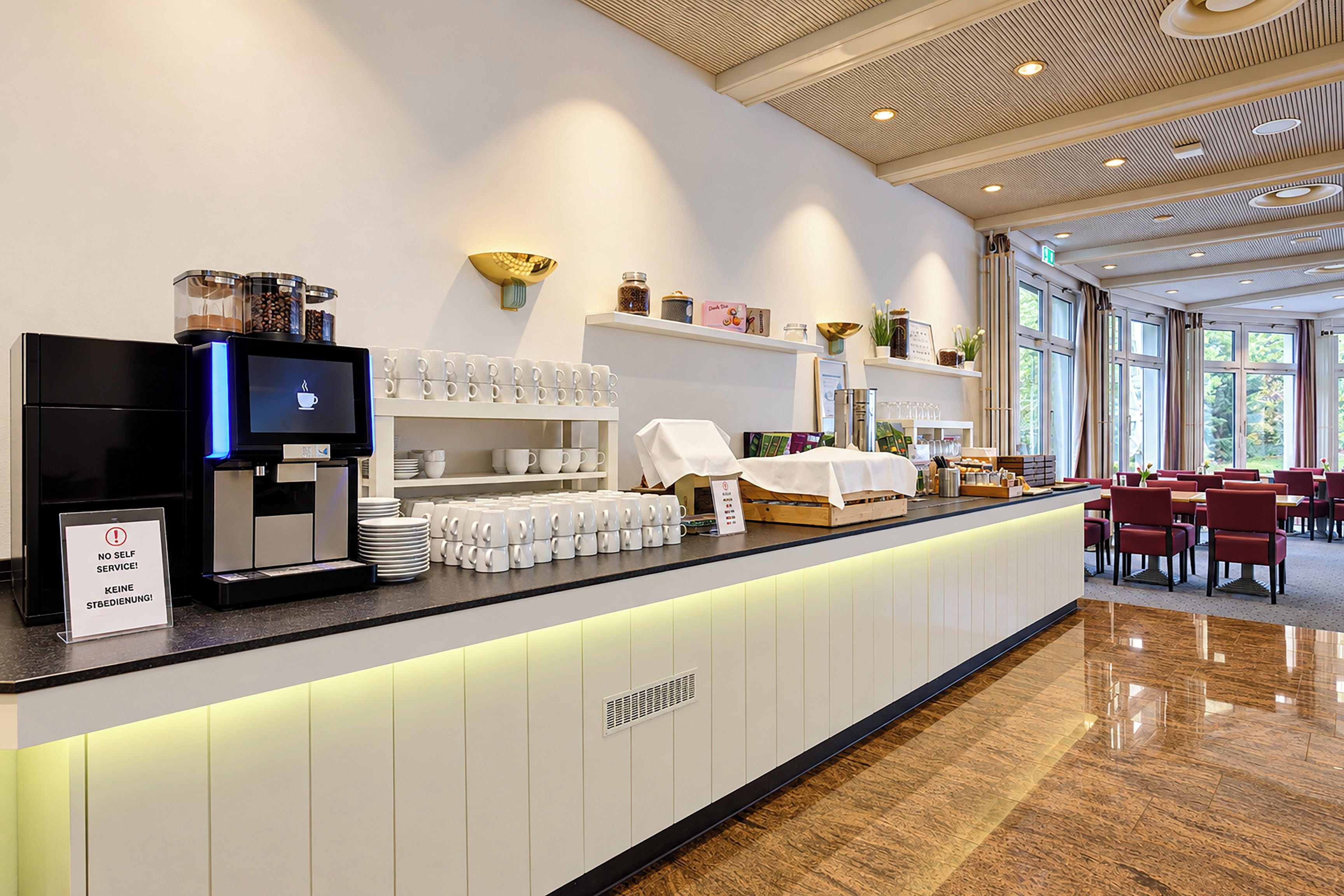 Una cafetería moderna con una máquina de café digital, una estación de autoservicio para café y un área de descanso con mesas y sillas.