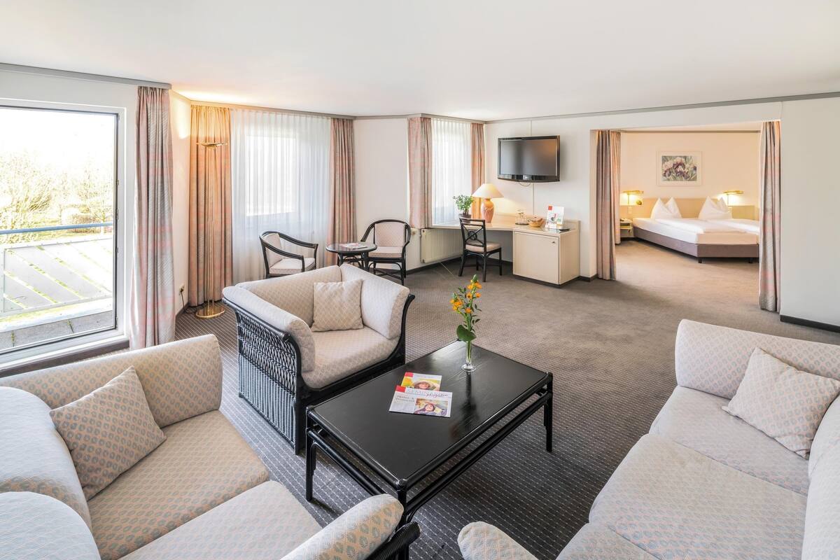 A hotel room with a sofa, armchair, dining table, TV, and a bed.