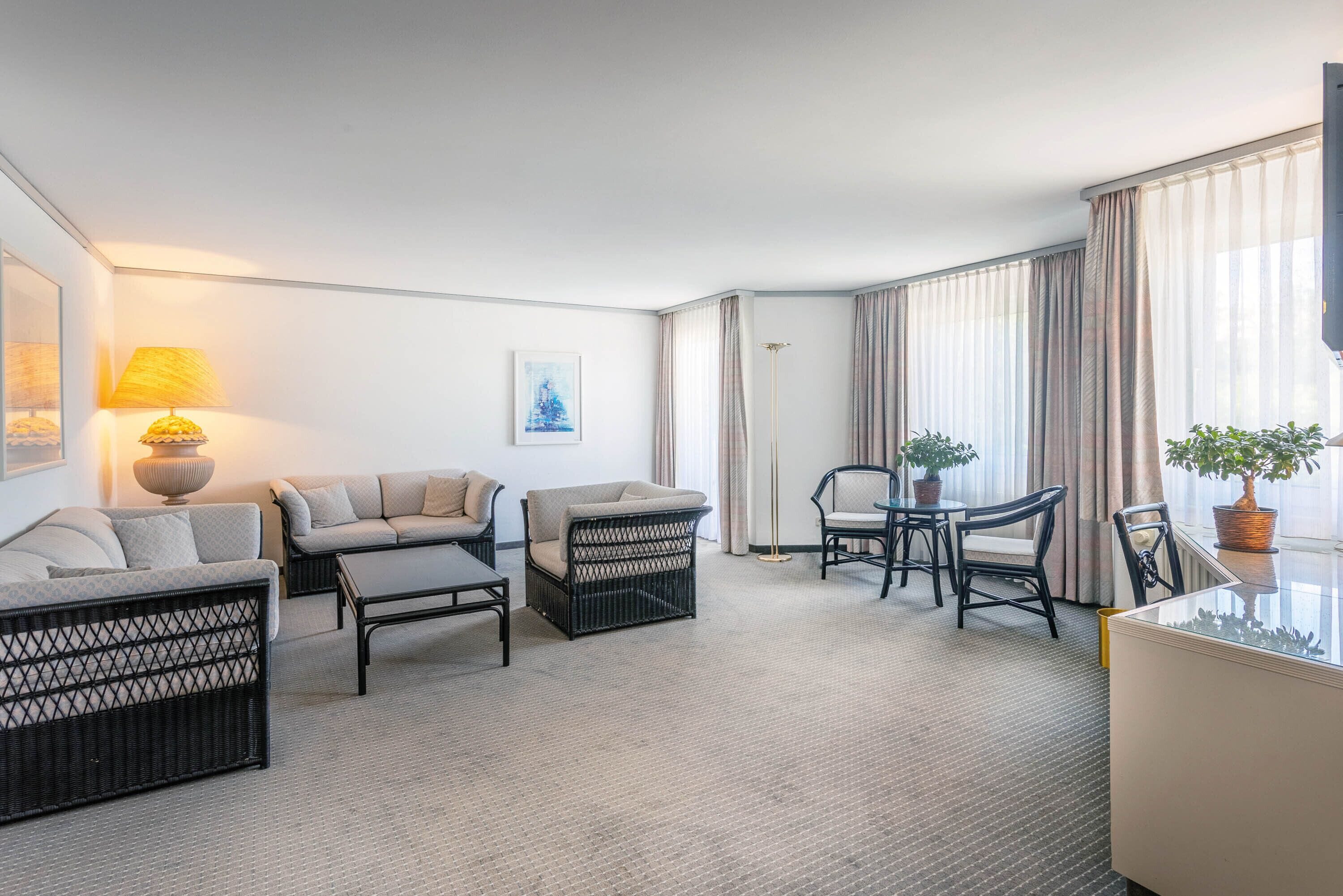 A modern hotel room with a sofa, armchair, and a small dining table with chairs. There is a floor lamp and a potted plant on the windowsill.