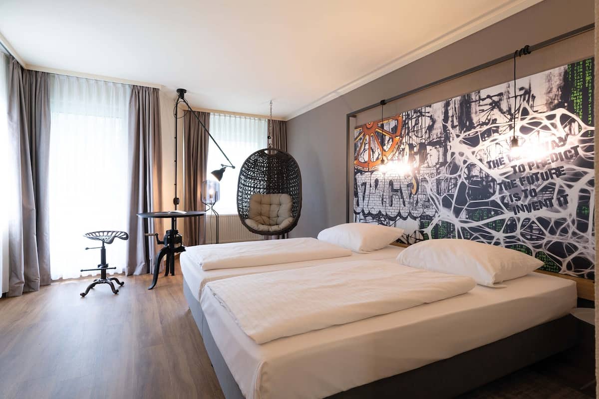 A modern hotel room with a large bed, a hanging chair, a desk, and a wall art piece.
