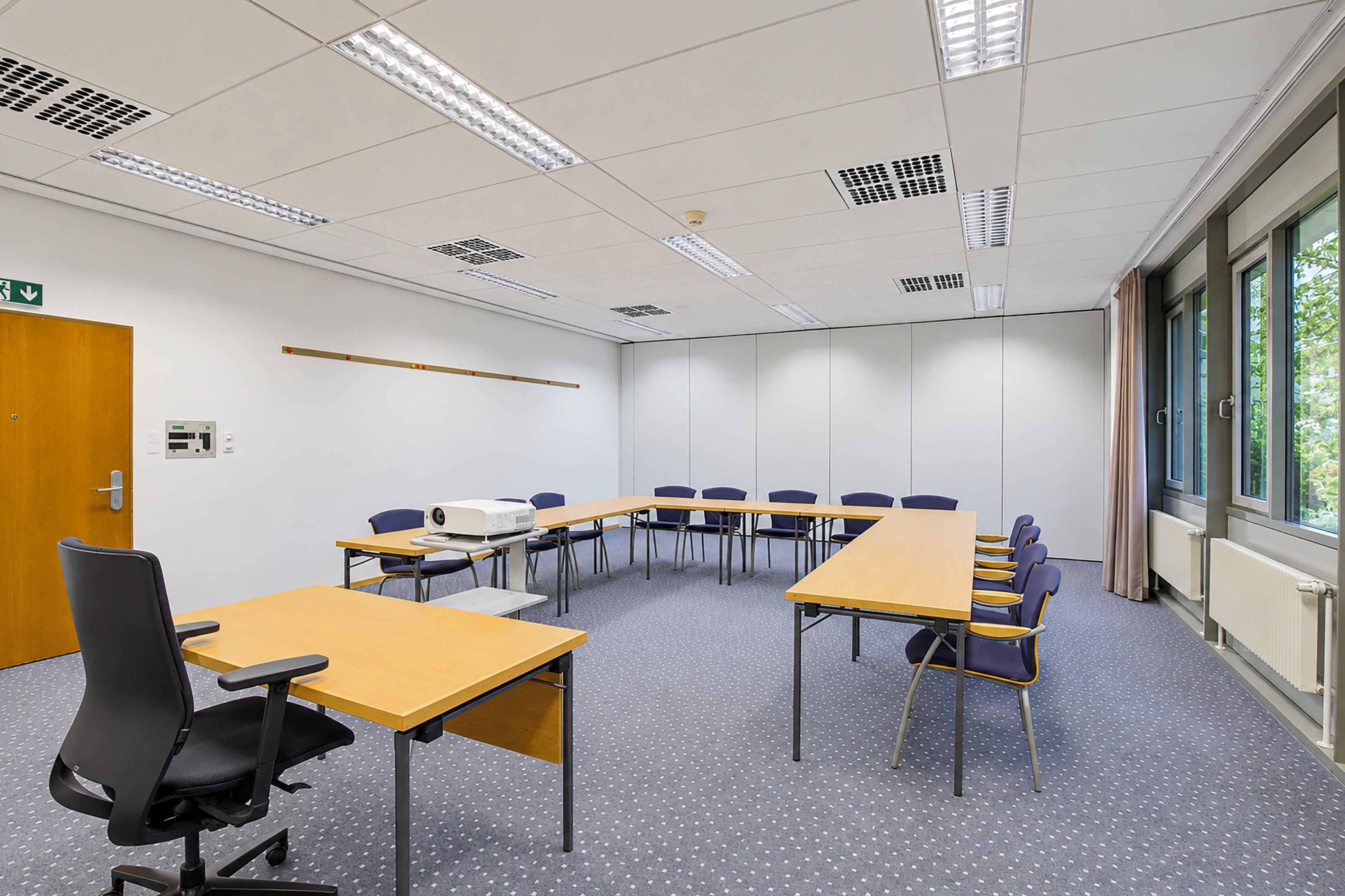 A conference room with a long table, chairs, a projector, and a door with an emergency exit sign.