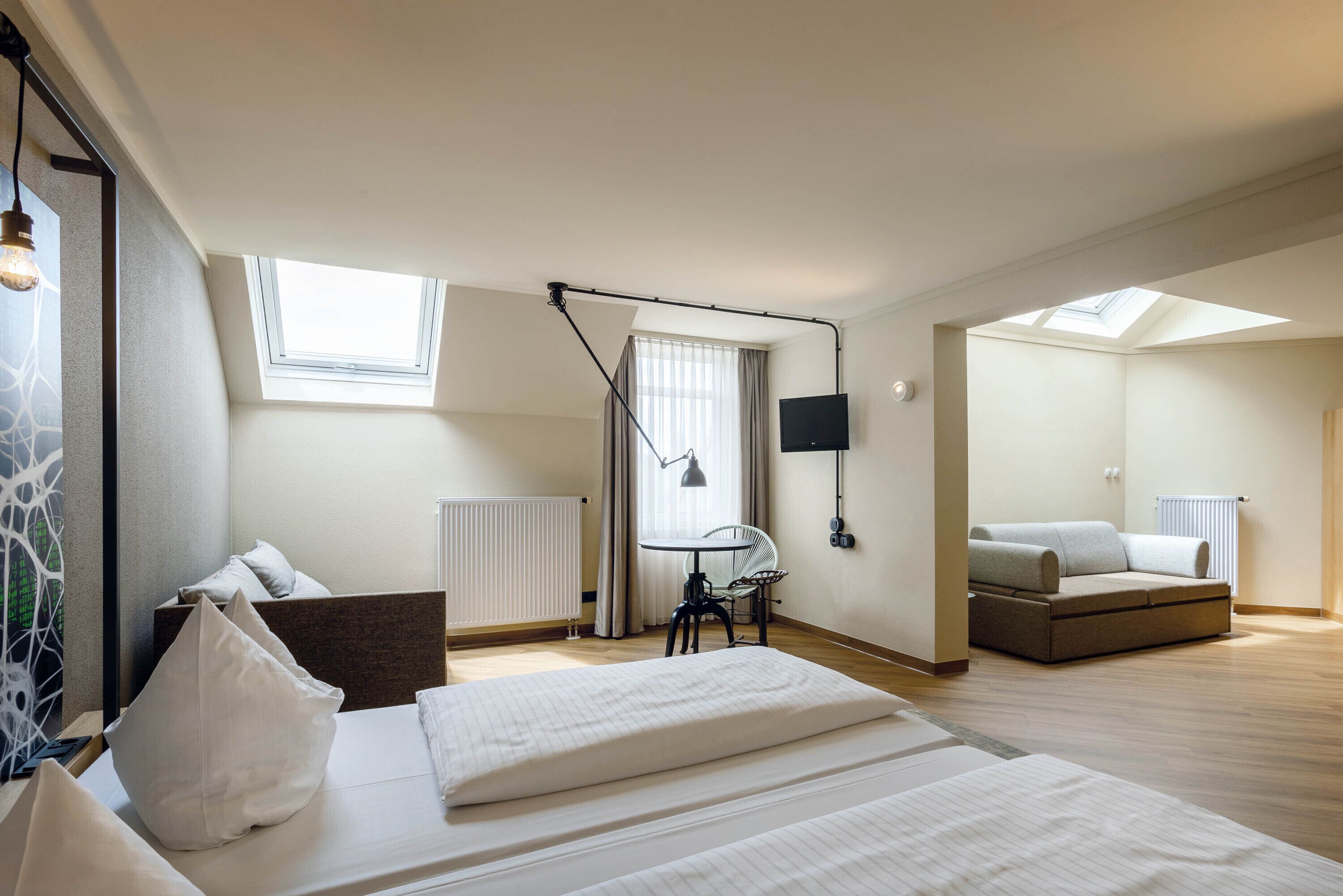 Une chambre moderne comprenant un lit, un bureau, une chaise, un canapé et un téléviseur. Elle est dotée de fenêtres de toit et d’une fenêtre qui laissent entrer la lumière naturelle.