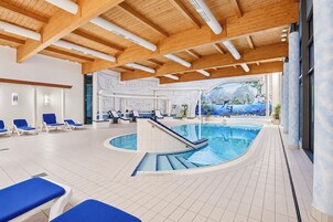 Piscina interior moderna com espreguiçadeiras azuis, teto de madeira e um mural com paisagem tropical.