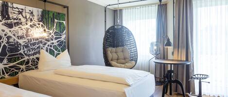 A hotel room with two beds, a hanging chair, a desk with a lamp, and a large abstract painting on the wall.