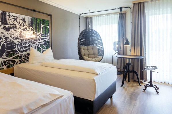 A hotel room with two beds, a hanging chair, a desk with a lamp, and a large abstract painting on the wall. - Seminaris Sunday Hotel Bad Boll (Bad Boll)