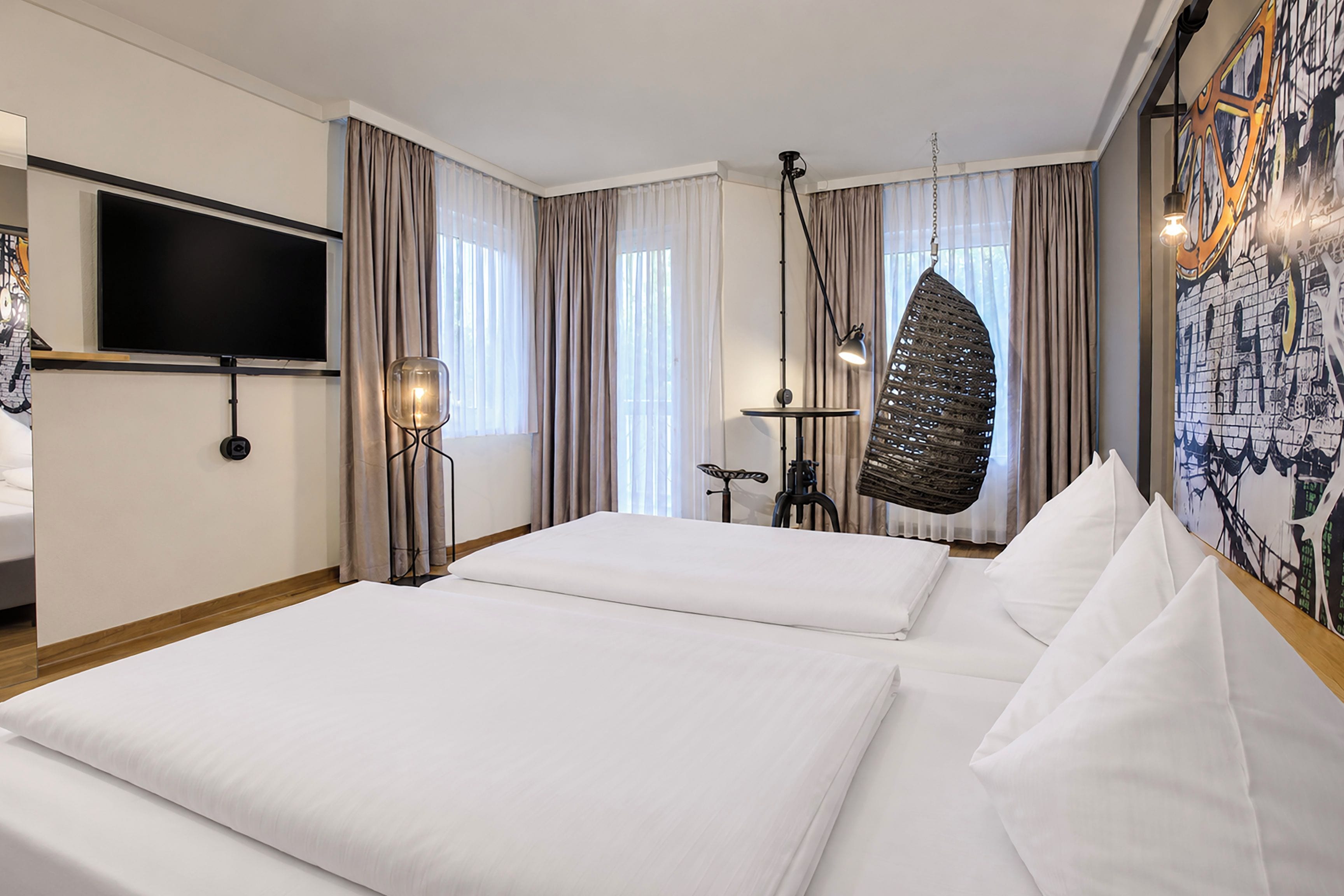 A hotel room with two beds, a flat-screen TV, a hanging chair, and a wall-mounted lamp.