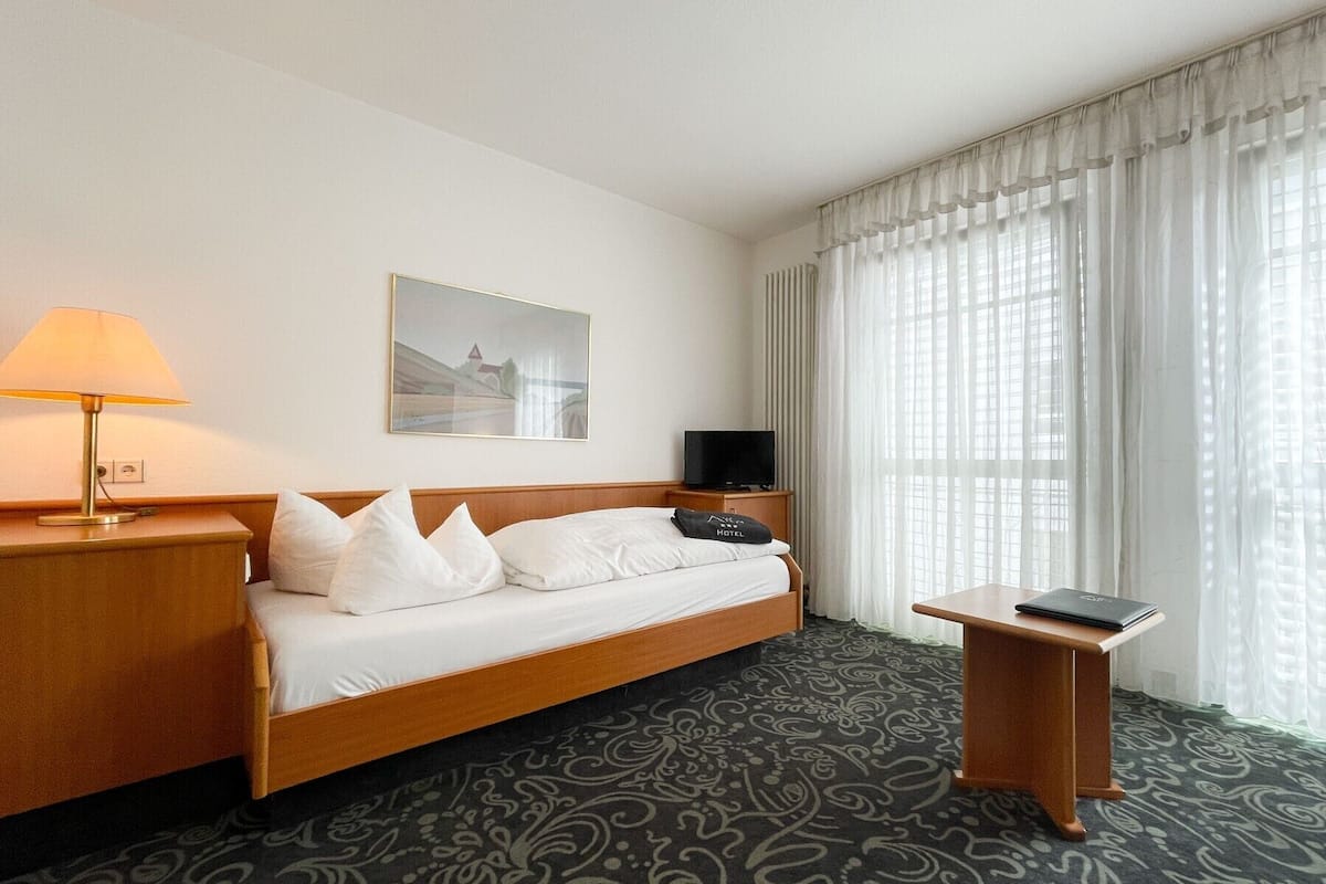 A hotel room with a bed, a bedside table, a lamp, a television, and a window with curtains.