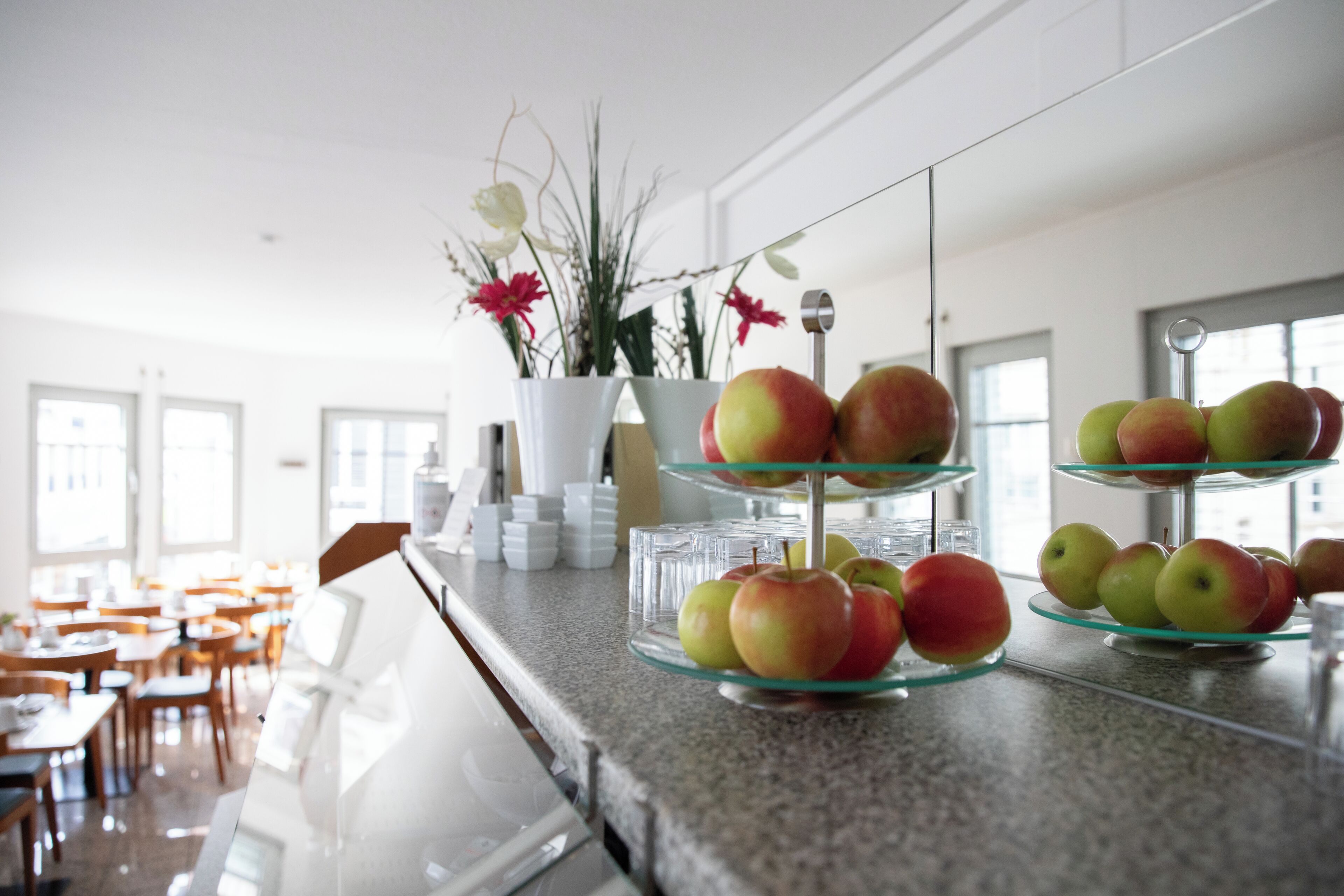 Zona de bufé con múltiples bandejas de vidrio que exhiben manzanas, un jarrón blanco con flores y un interior moderno y luminoso con luz natural.