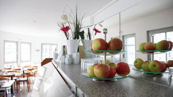 Zona de bufé con múltiples bandejas de vidrio que exhiben manzanas, un jarrón blanco con flores y un interior moderno y luminoso con luz natural.