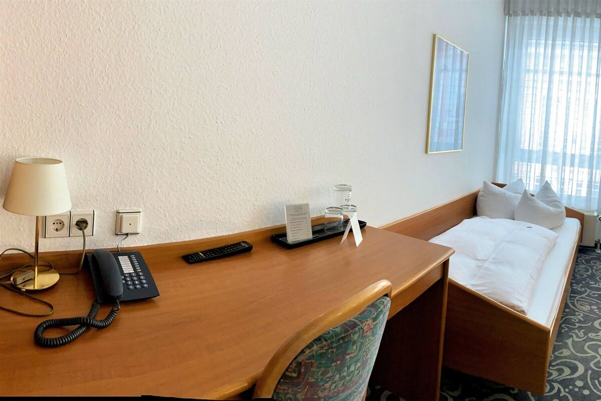 A hotel room with a desk, two chairs, a bed, and a large window with curtains.