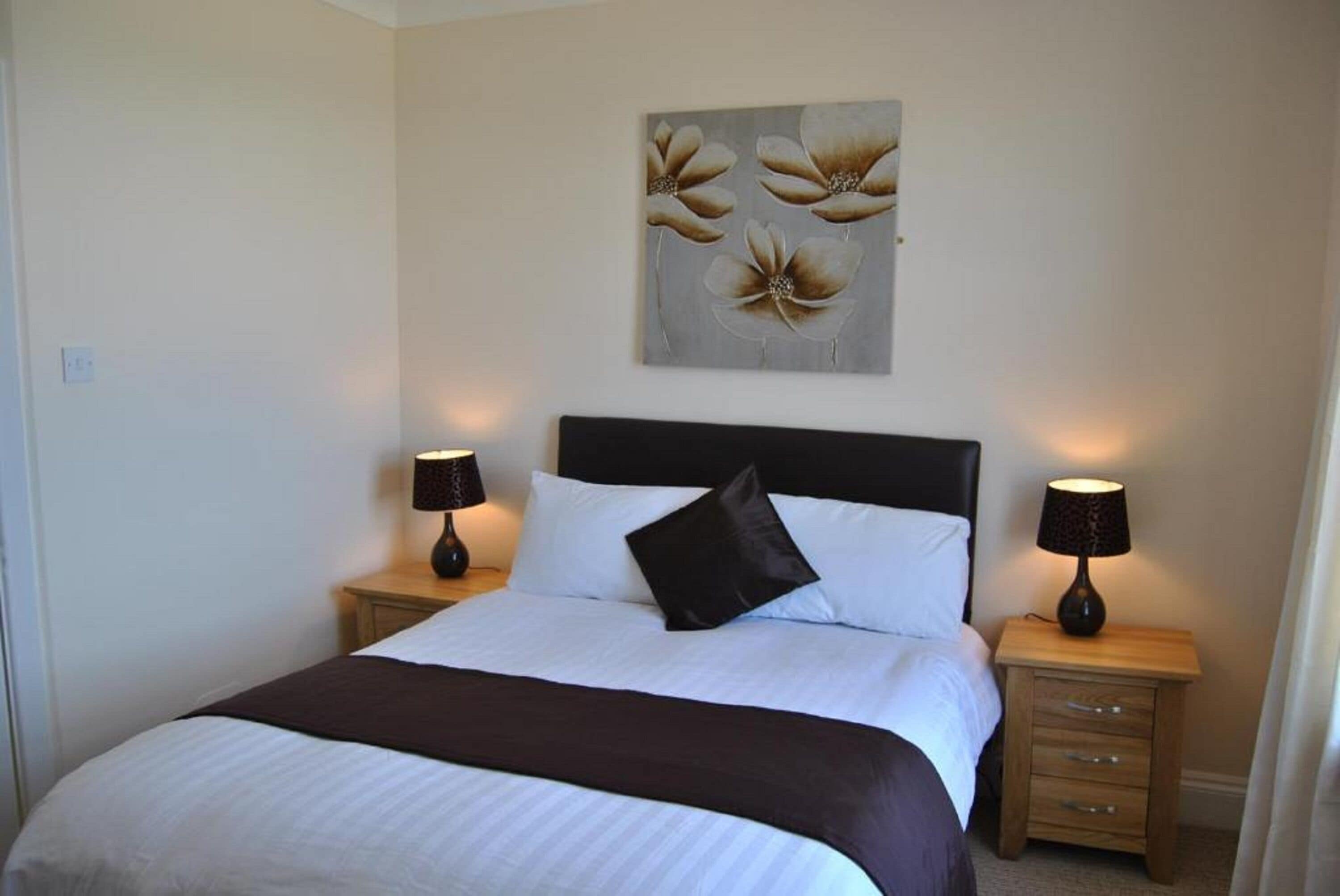 Uma cama bem arrumada com travesseiro preto, duas mesas de cabeceira com abajures e uma obra de arte floral sobre a cama.