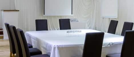 A conference room with a large oval table, black chairs, white curtains, and a projector screen.
