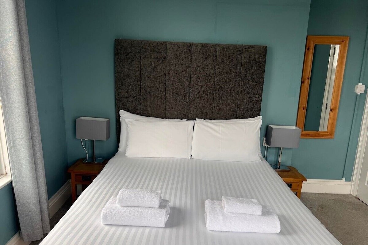 A neatly made bed with white linens and two folded towels, a dark headboard, two bedside tables with lamps, and a mirror on the wall.