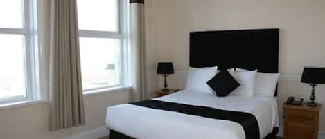 Una habitación de hotel con una cama grande, dos mesitas de noche, un ventanal con cortinas y una pared lisa.
